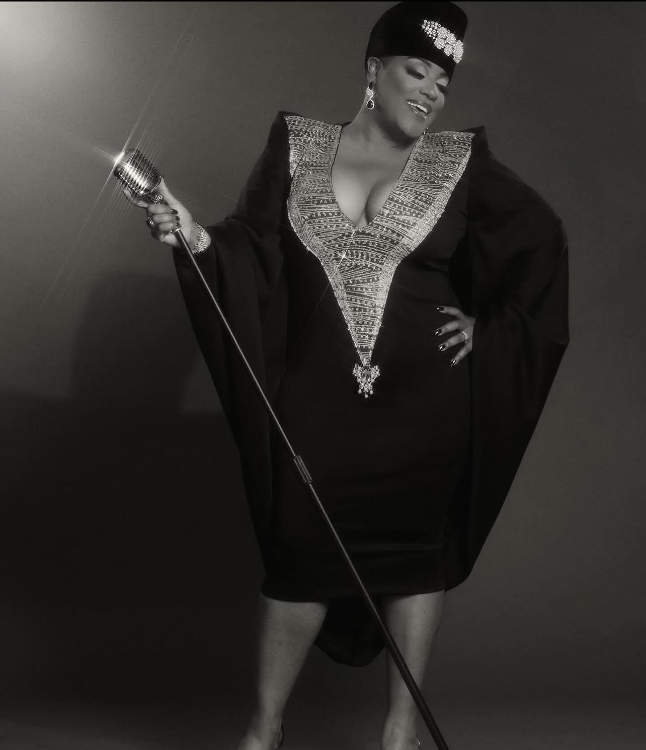 Woman in elegant dress holding a microphone, smiling, with a hand on her hip, in a studio setting.