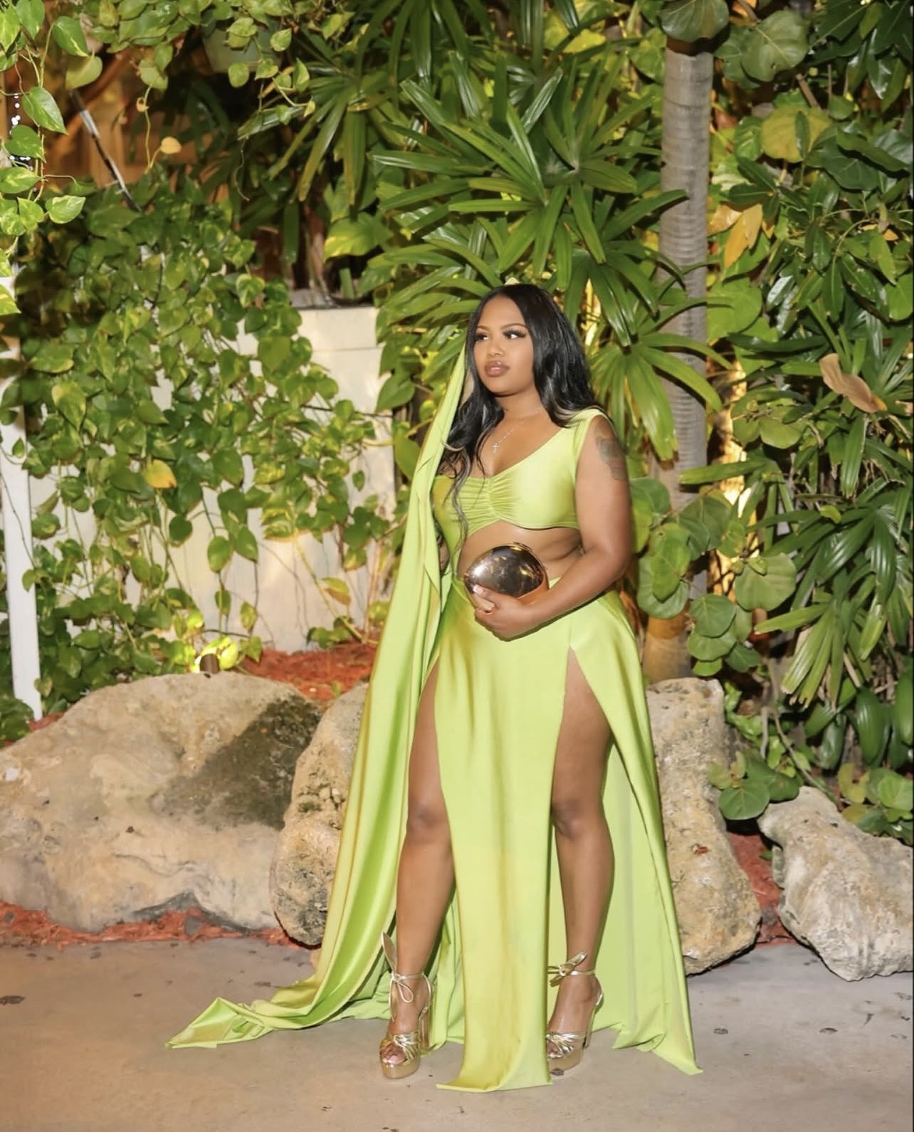 Woman in lime green dress holding a clutch, standing outdoors among plants and rocks.