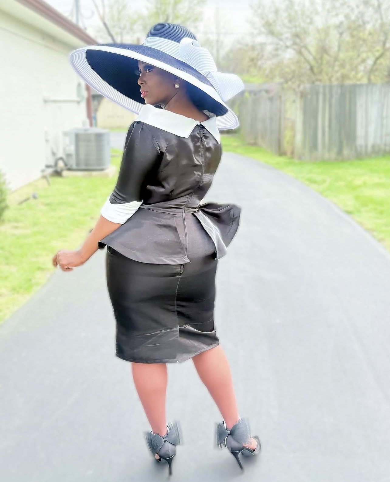 Woman in black dress and large white and blue hat walking on a paved path, with a fence and trees in background.
