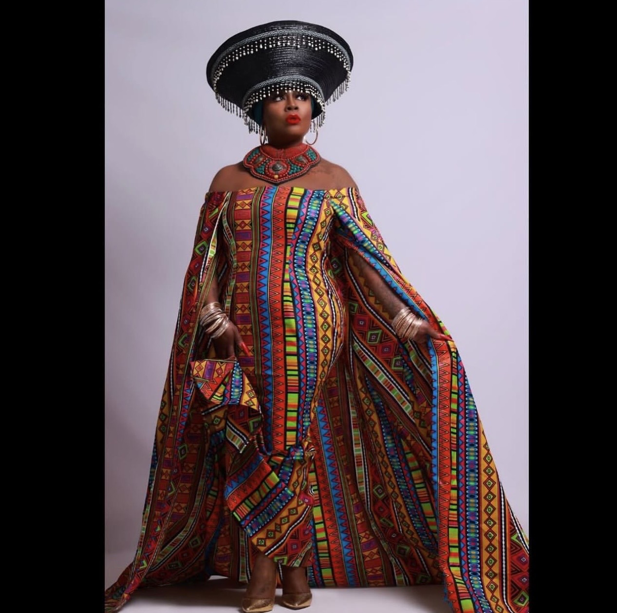 Woman in colorful traditional attire with a large black headpiece, standing against a plain background.