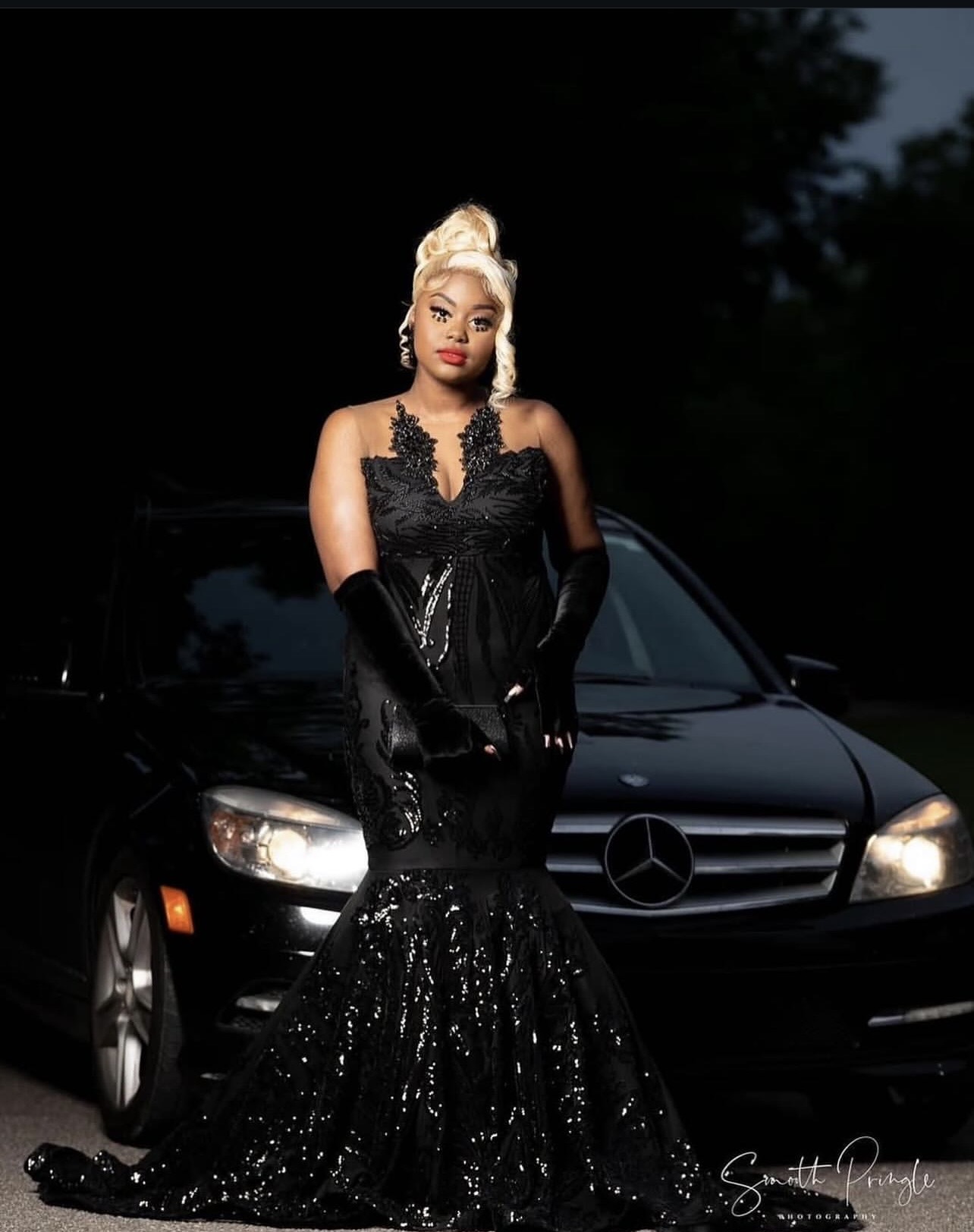 Woman in black gown and gloves standing in front of a black Mercedes-Benz car outdoors at night.