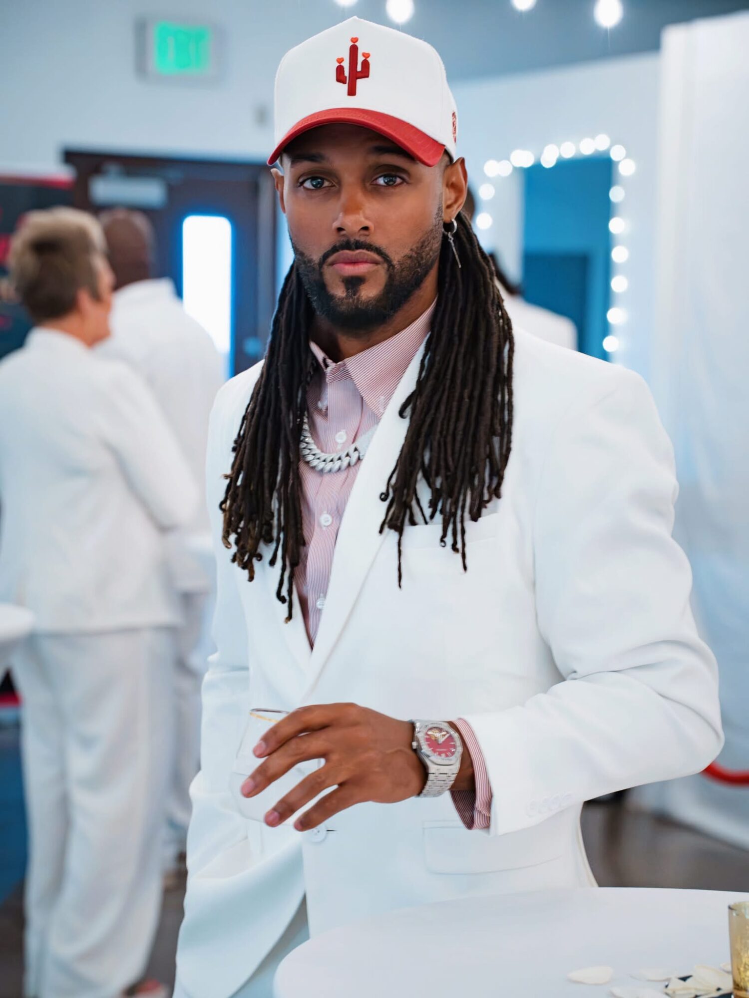 Man with long dreadlocks wearing a white suit, red and white cap, and jewelry, standing indoors.