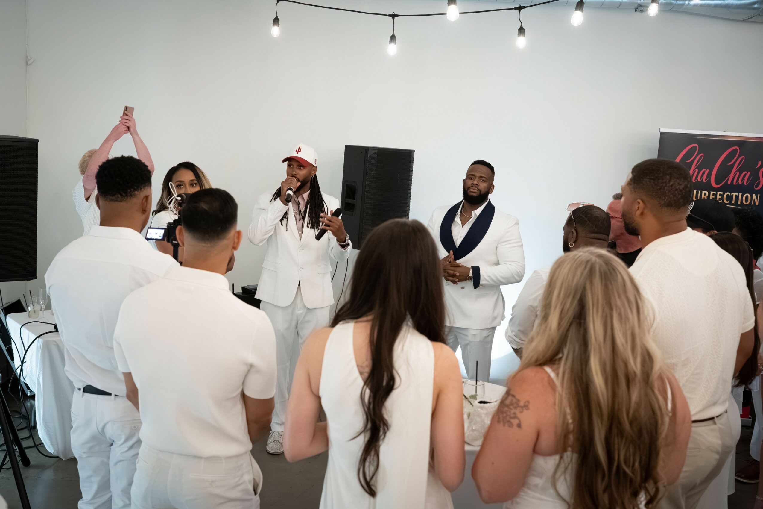 Group of people gathered indoors, some singing or speaking, with a white wall and string lights overhead.