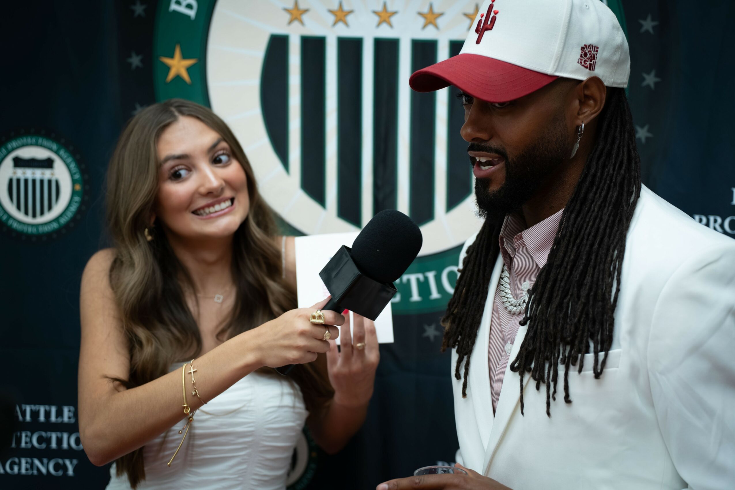 Young woman interviewing a man with a microphone, both smiling, in front of a sports team logo backdrop.