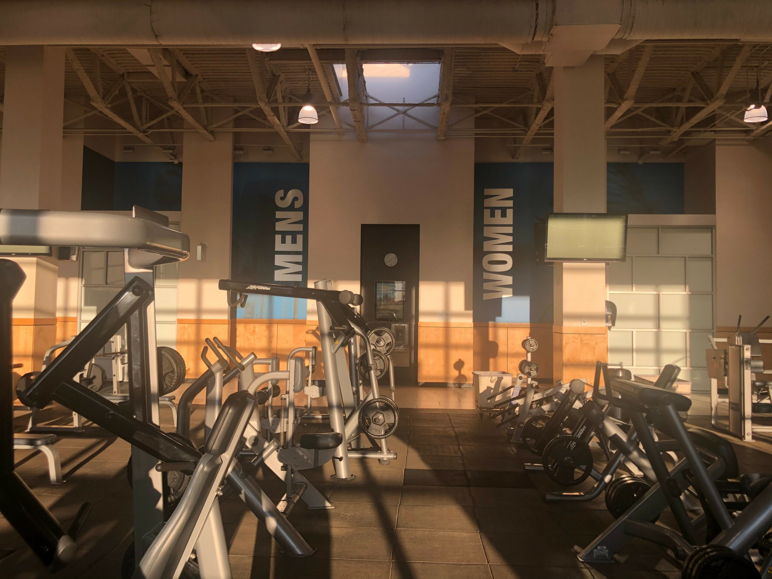 Gym interior with exercise equipment, signs for men's and women's restrooms, and a window in the background.