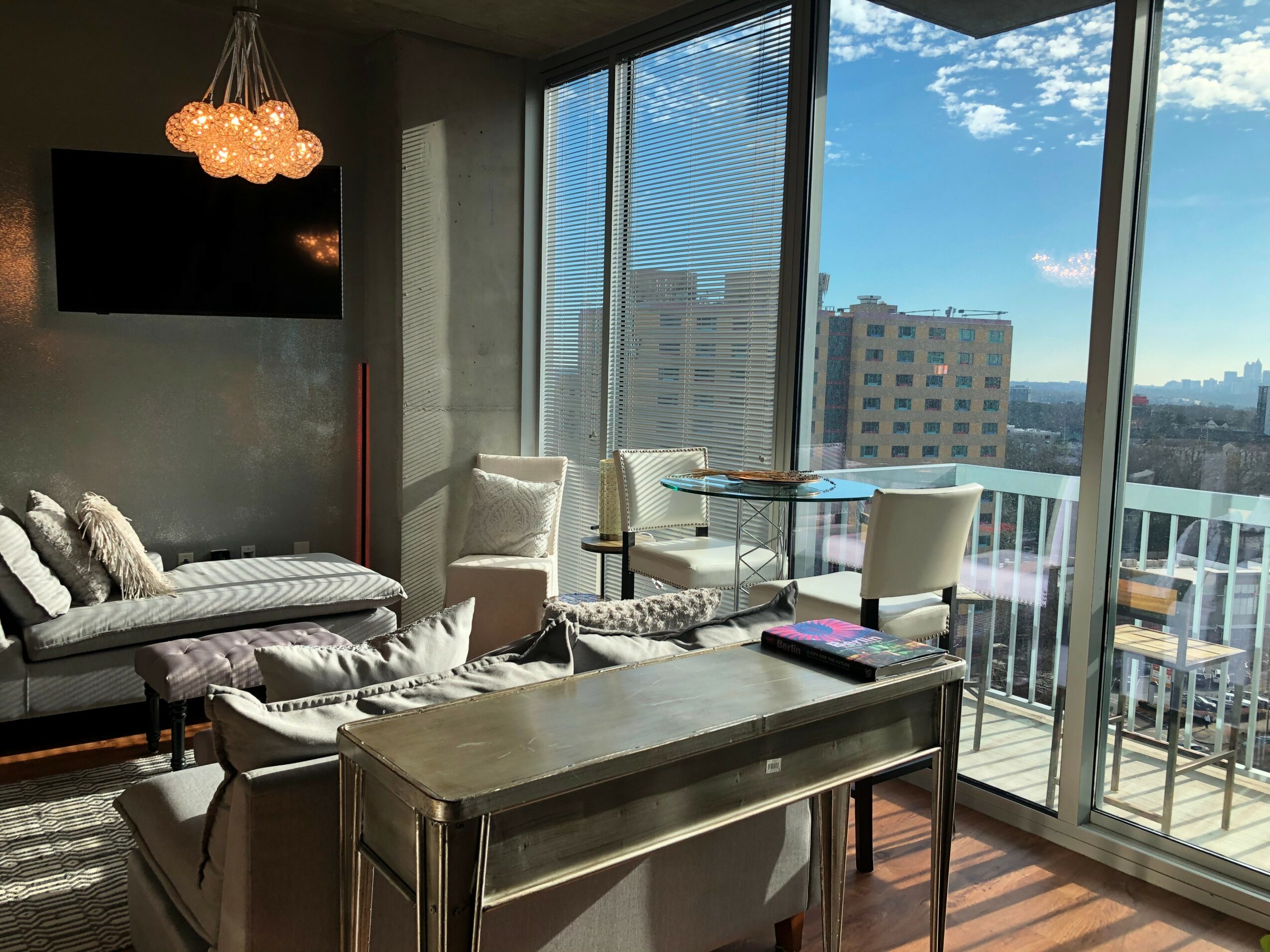 Living room with sofa, chairs, TV, and large windows showing city buildings and blue sky.