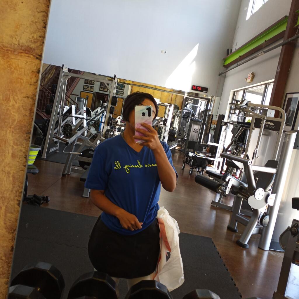 Person taking a mirror selfie in a gym, wearing a blue shirt and black pants, with gym equipment in background.