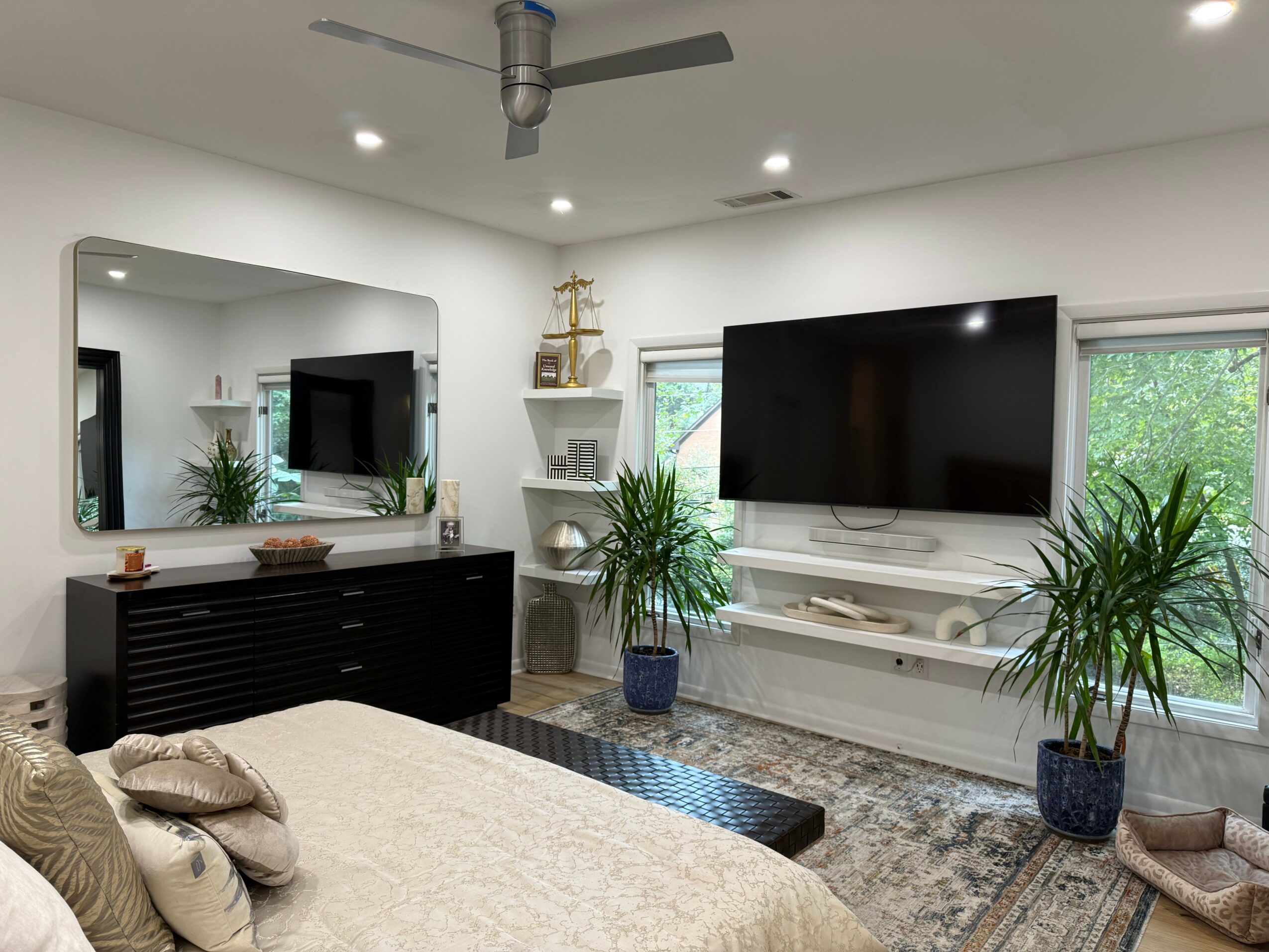Living room with large wall mirror, flat-screen TV, plants, and a bed with pillows, bright windows, and modern decor.