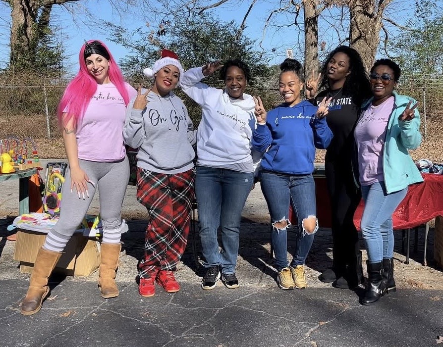 Six women standing outdoors, smiling, with trees in the background, some wearing casual clothing and holiday accessories.