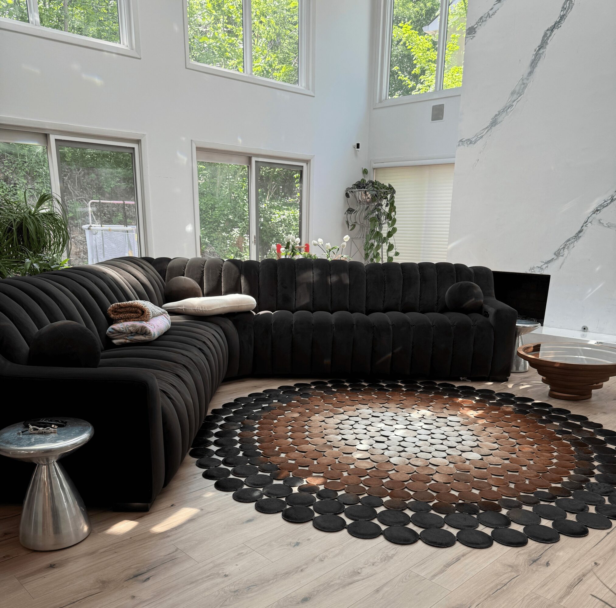 Living room with large black sectional sofa, round patterned rug, and tall windows with greenery outside.