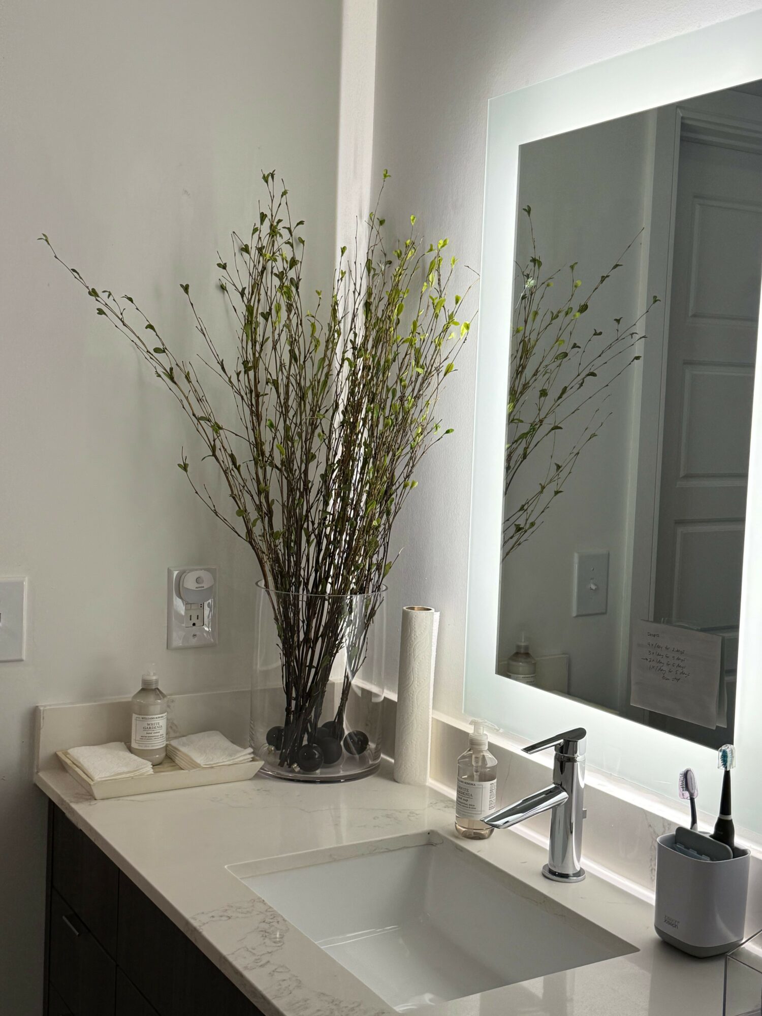 Bathroom sink with a large vase of branches, a soap dispenser, and candles on the countertop.