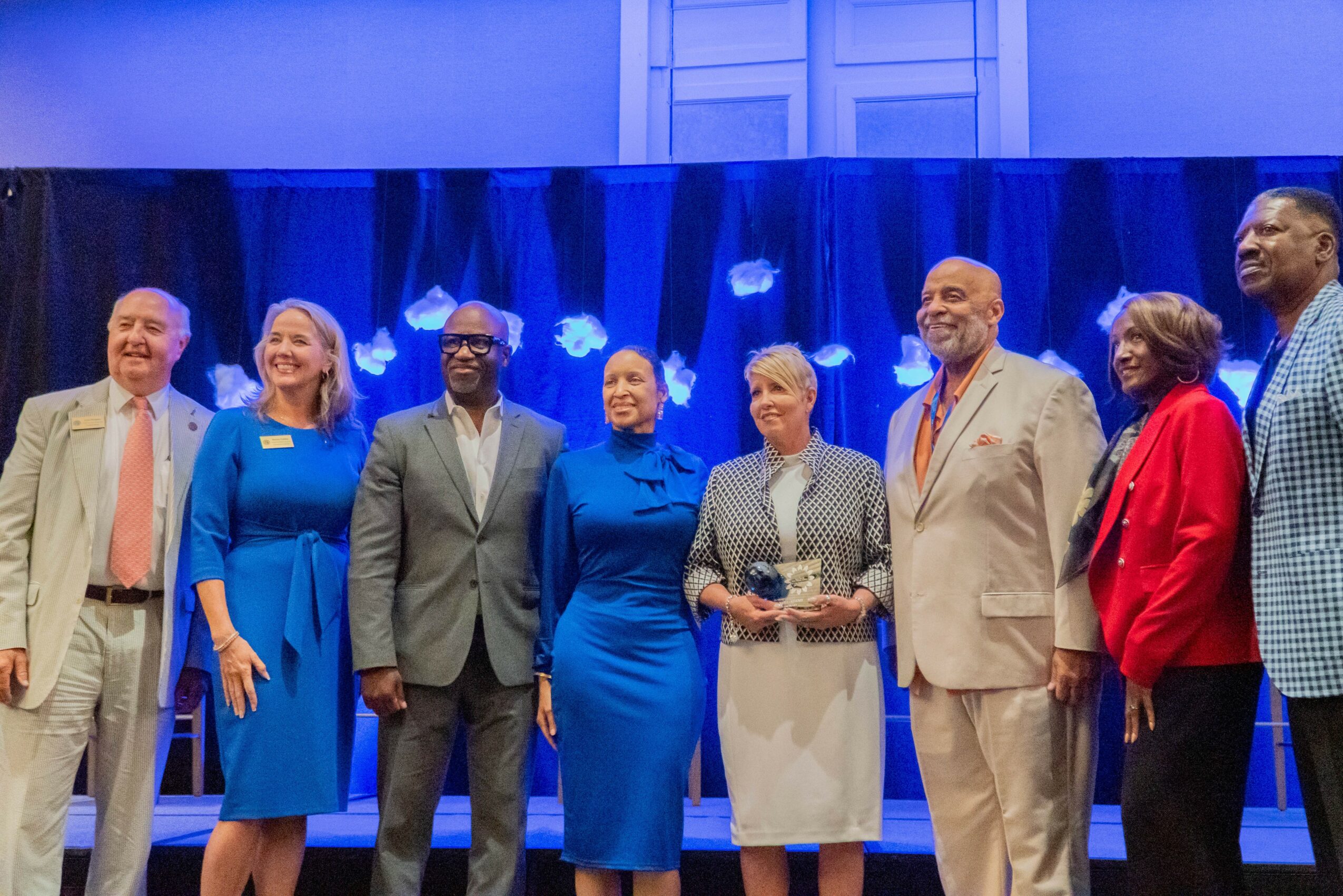Group of seven people standing on stage, smiling, with a blue curtain background.