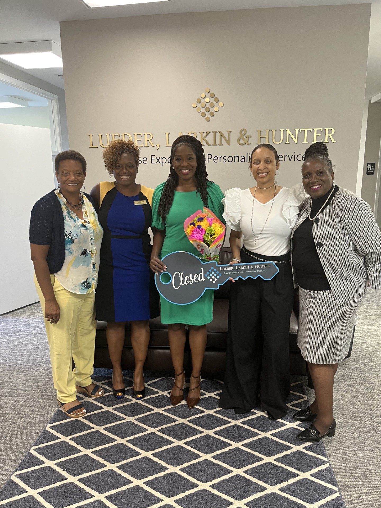 Five women standing together indoors, one holding a bouquet and a large key-shaped sign, smiling for the photo.