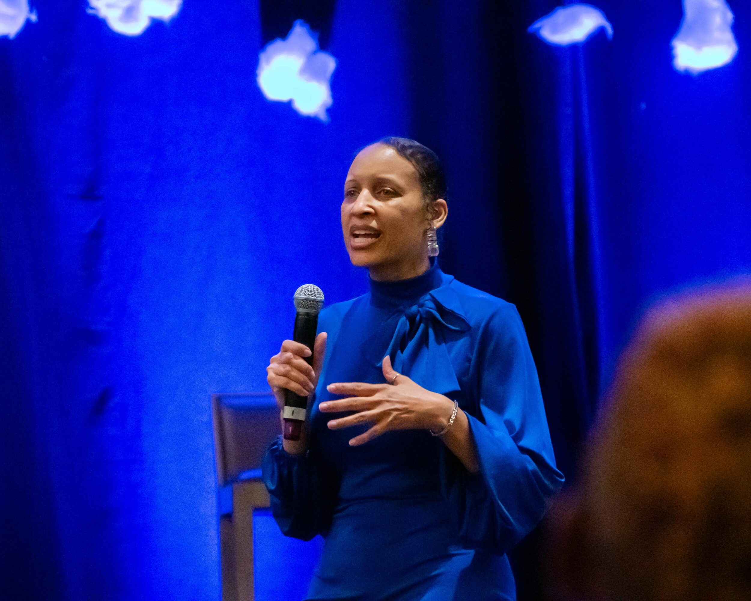 Woman speaking into microphone on stage with blue background and hanging cloud decorations.