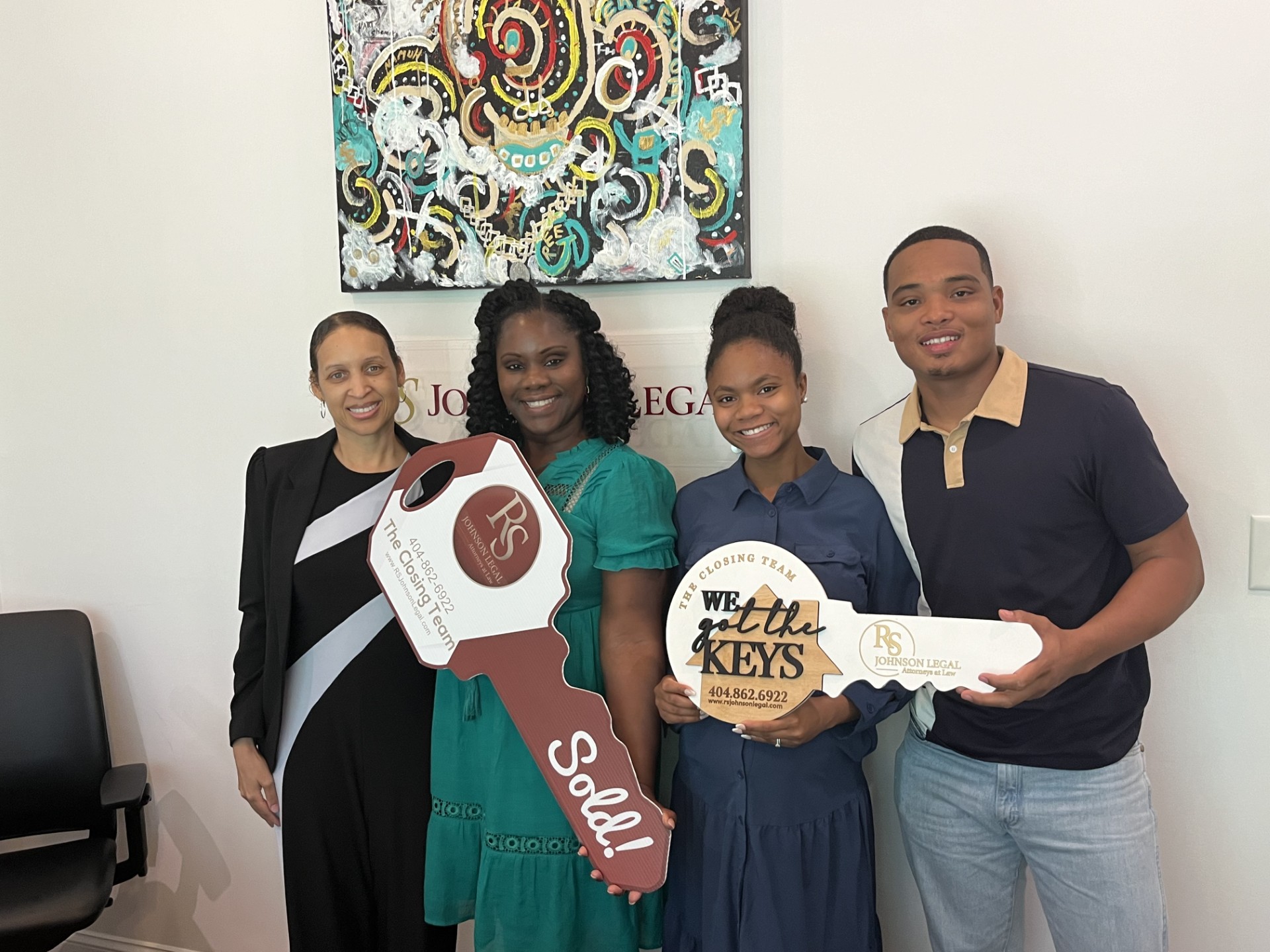 Four people standing together indoors, smiling, holding a large key-shaped sign and a circular sign, with a colorful abstract painting behind them.