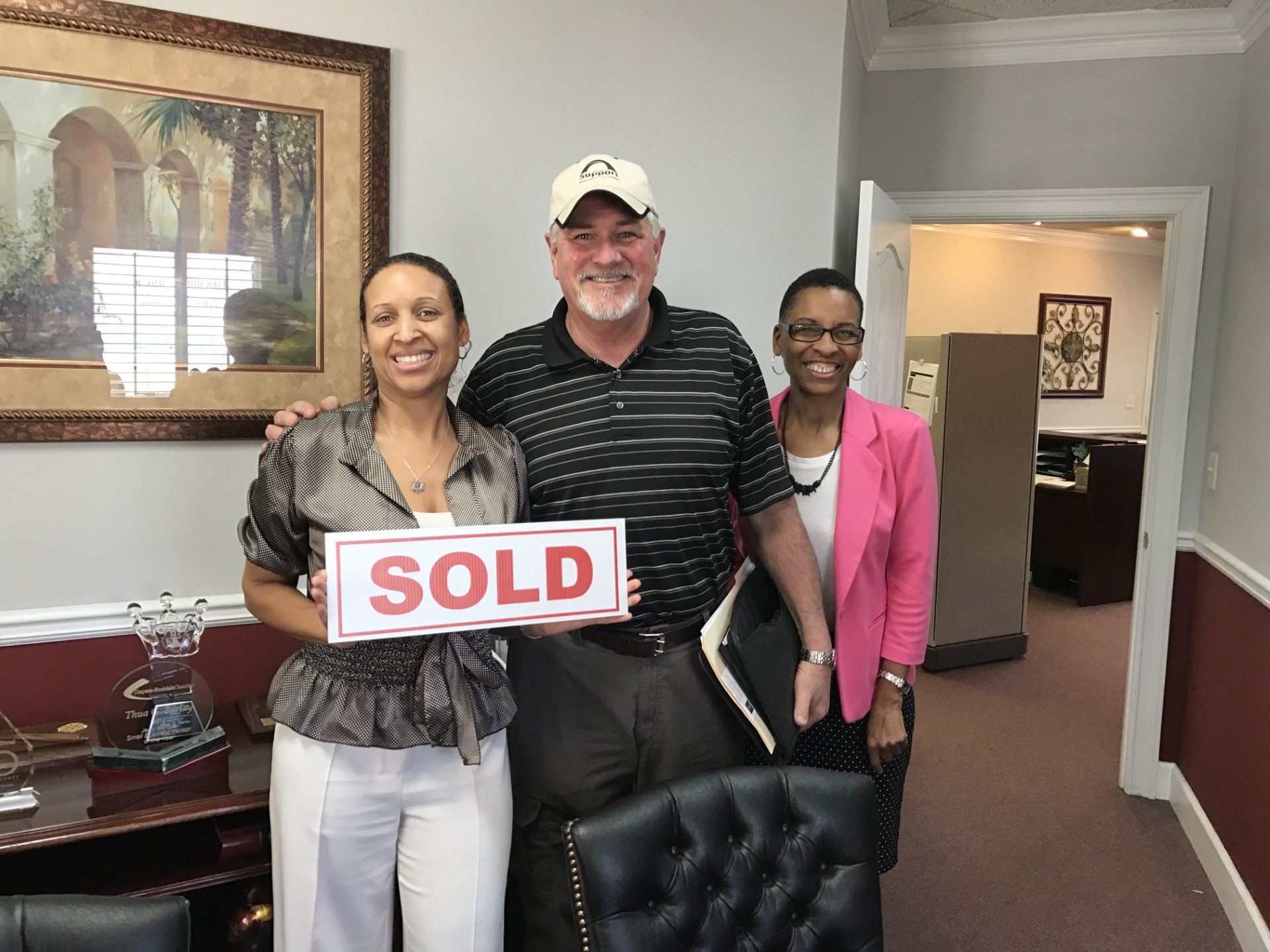 Three people smiling indoors, one holding a 'SOLD' sign, in a room with artwork and furniture.