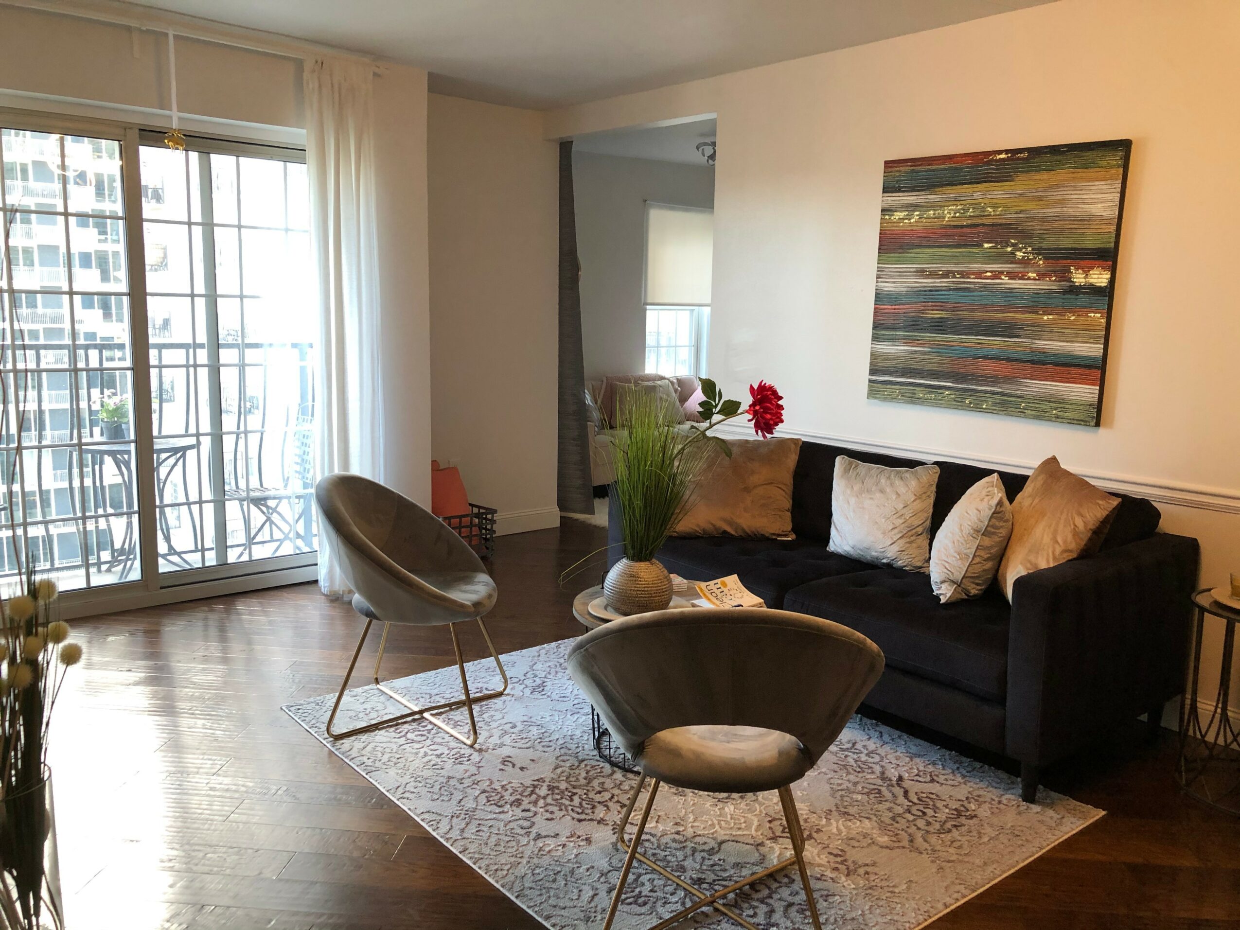 Living room with a black sofa, two chairs, a rug, and a large window with balcony doors, sunlight streaming in.