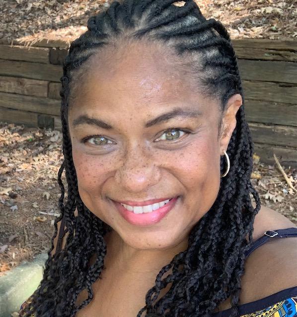 Smiling woman with braided hair outdoors, wearing hoop earrings and a dark top, in front of a wooden background.