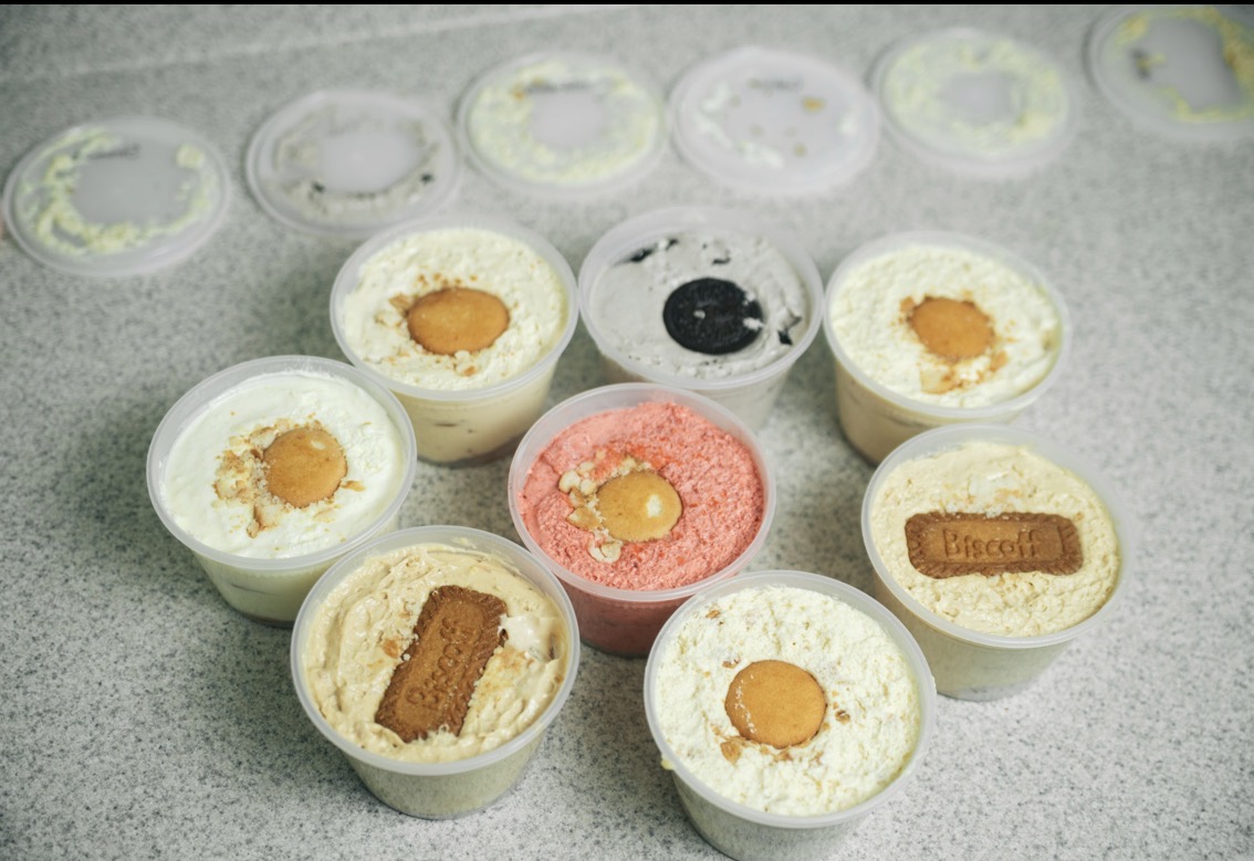Six small cups of dessert with various toppings and a pink one in the center, on a gray surface.