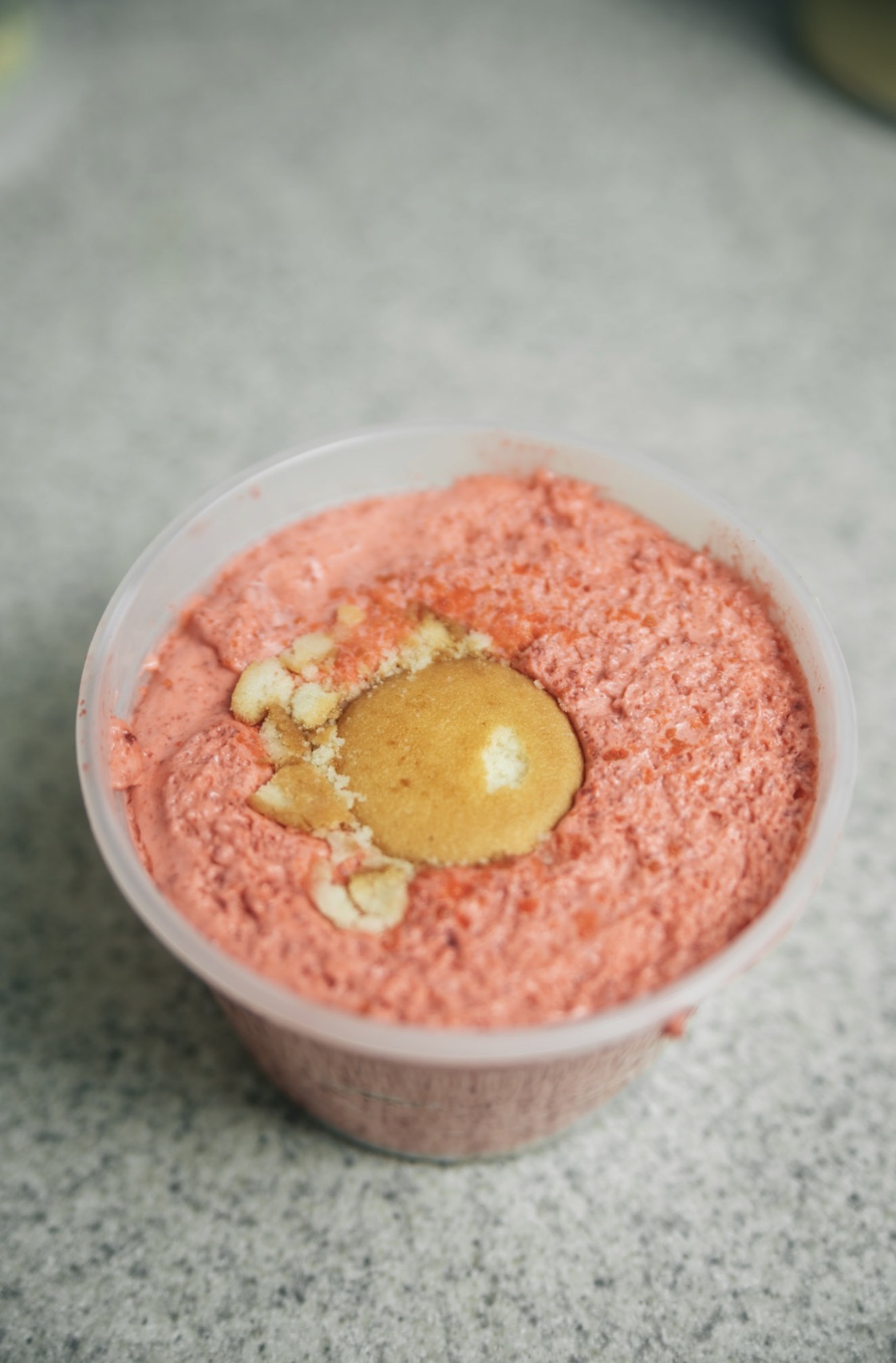 Pink frozen dessert with a dollop of yellow sauce and small white chunks in a clear cup on a gray surface.