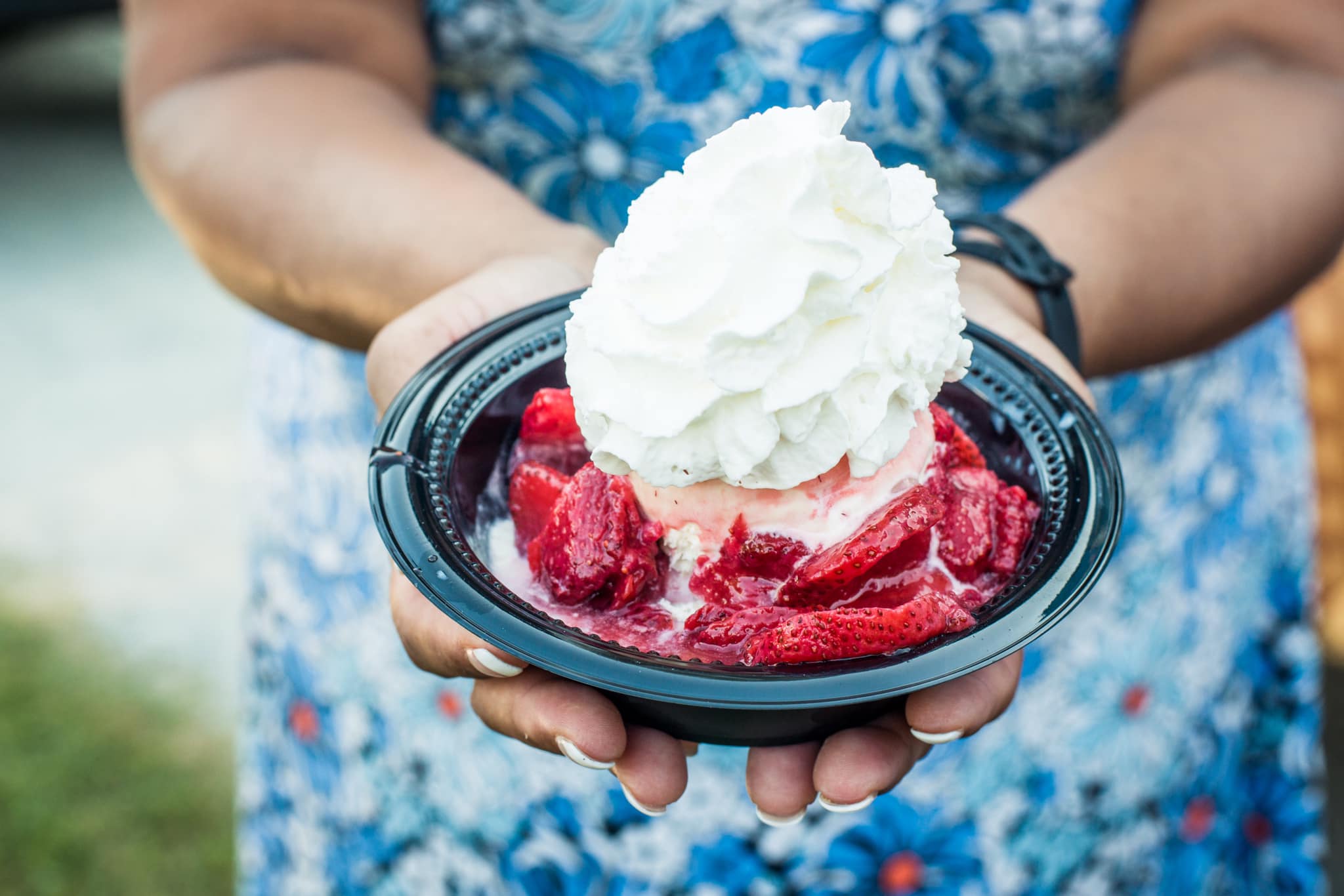 Person holding a black bowl with whipped cream and red fruit, wearing a blue floral shirt.