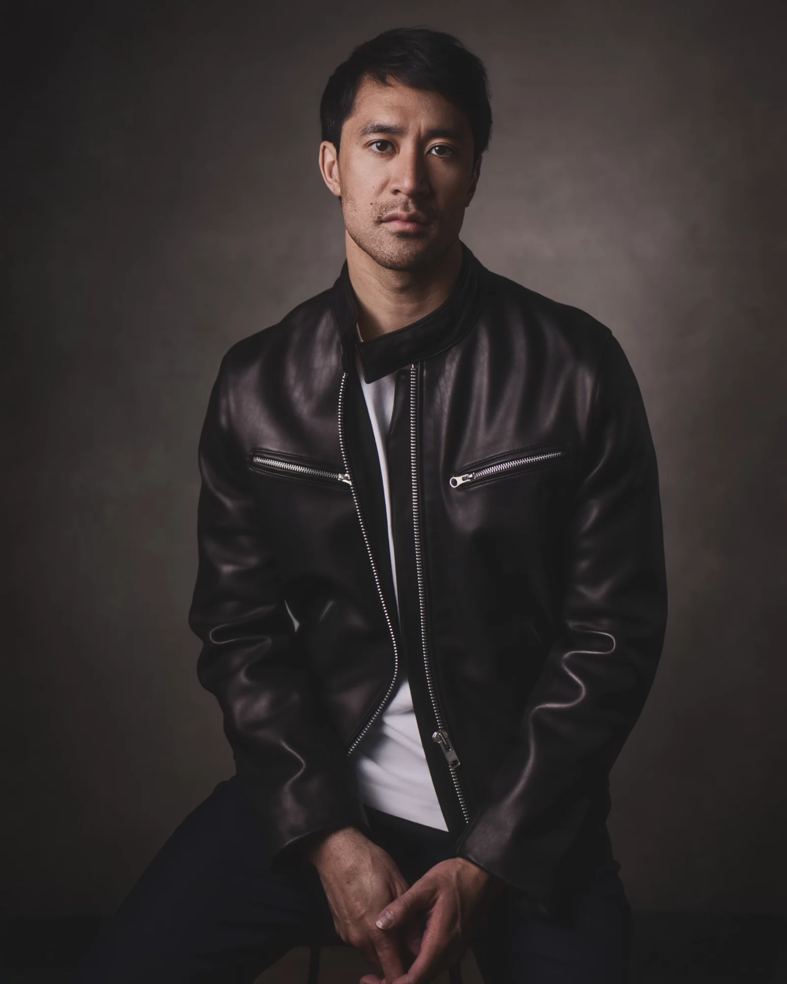Man with short dark hair wearing a black leather jacket and white shirt, standing against a dark background.