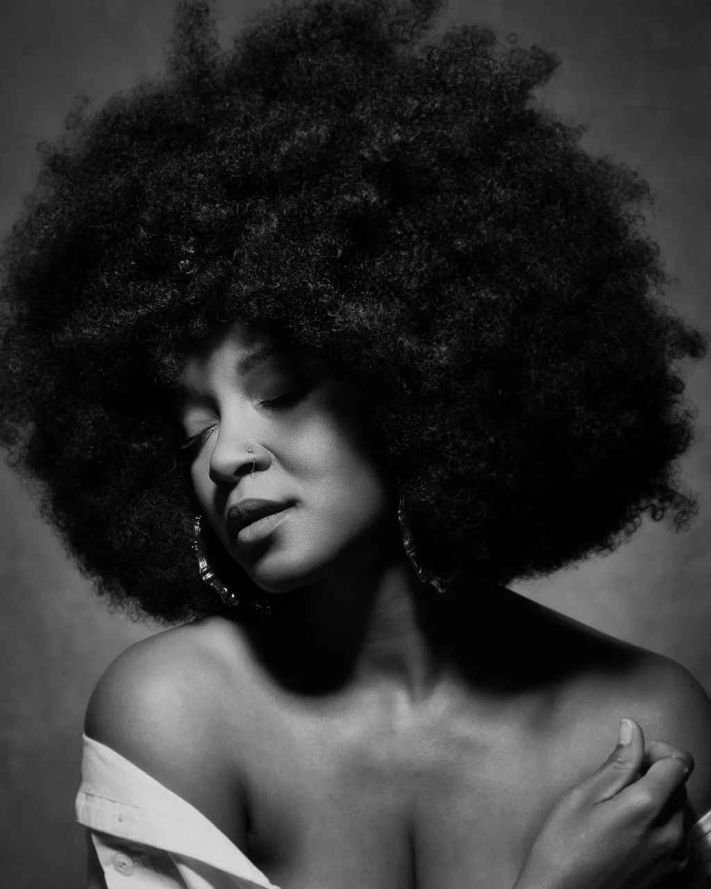 Black and white portrait of a woman with large, curly hair, eyes closed, and one hand on her shoulder.