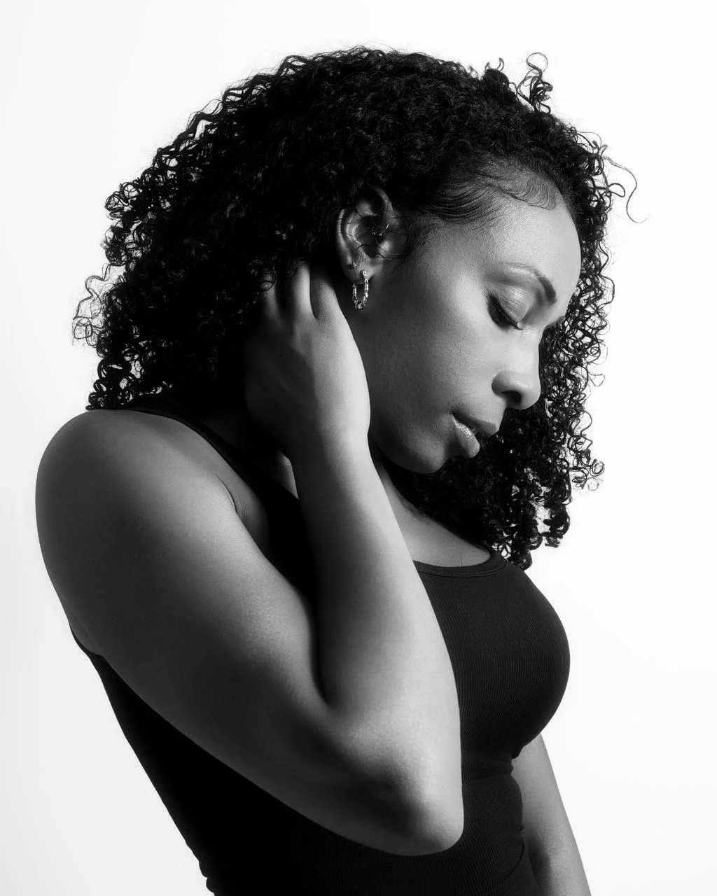 Black and white portrait of a woman with curly hair, eyes closed, touching her neck, wearing a sleeveless top.