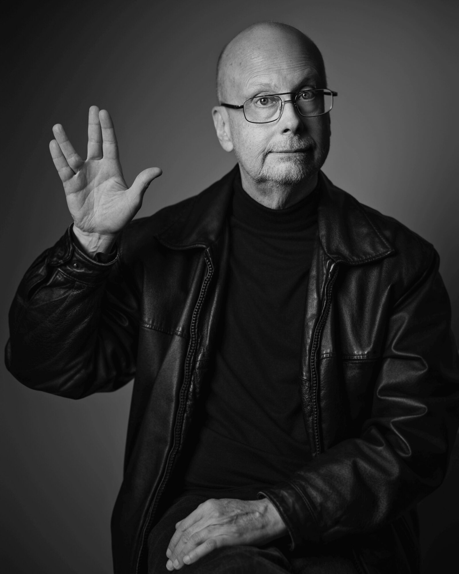 Bald man with glasses wearing a leather jacket, raising his right hand, in black and white.