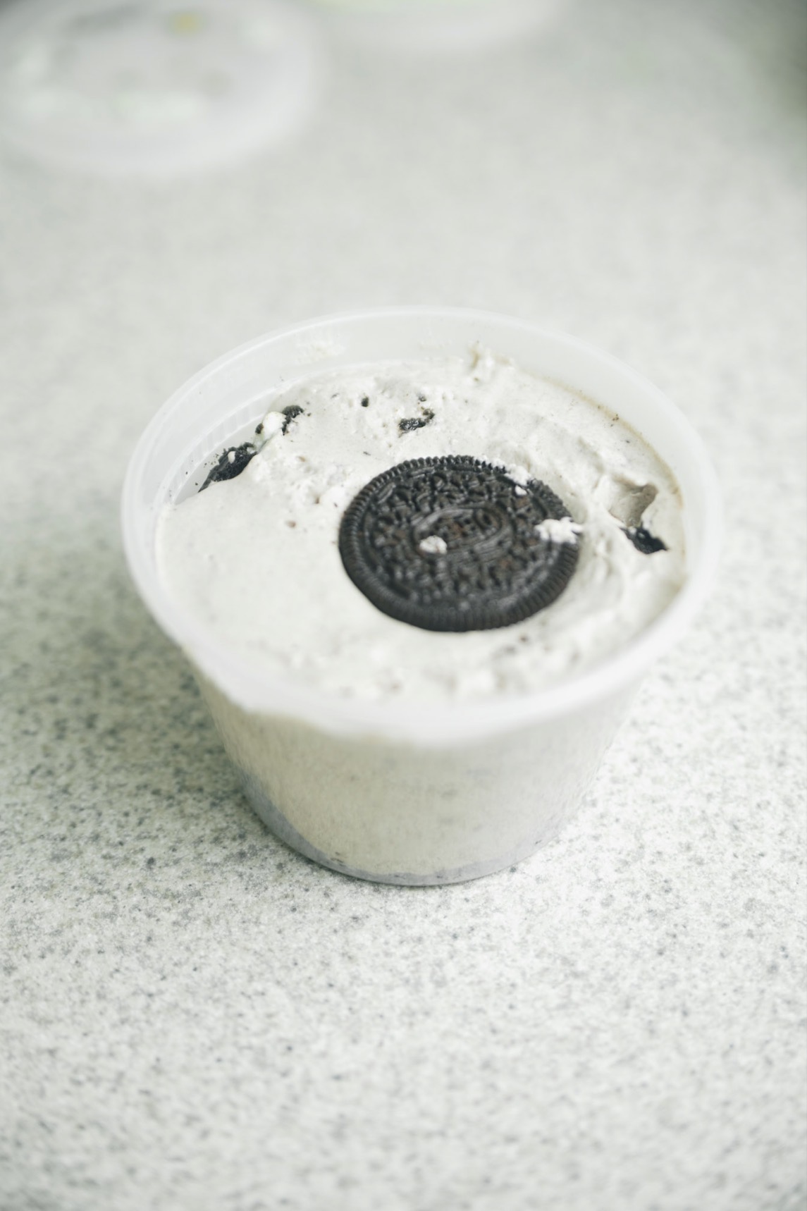 Container of cookies and cream ice cream with an Oreo on top, on a light-colored surface.