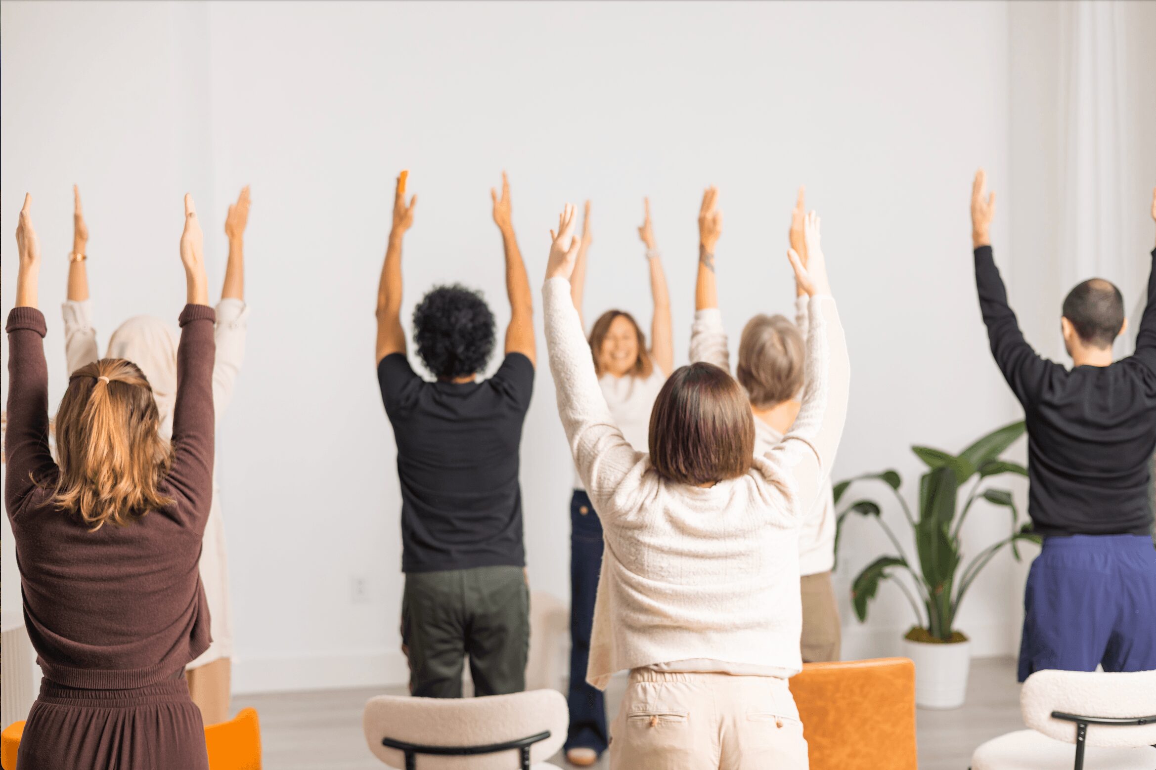 Group of people raising their hands in a room with white walls and potted plants, facing forward.