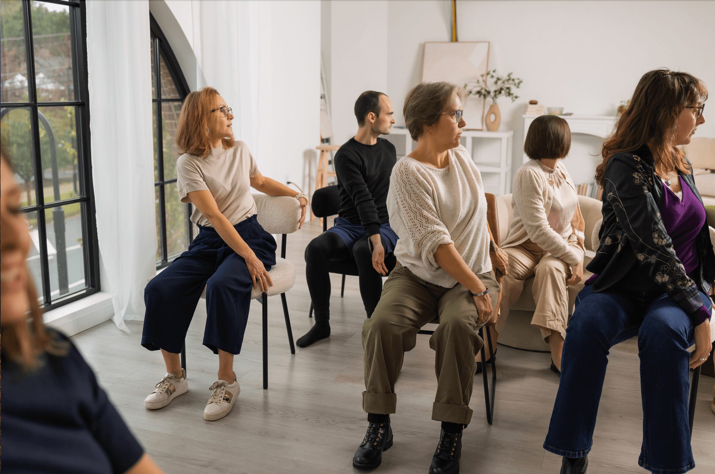 Group of women sitting on chairs in a bright room with large windows, participating in a class or workshop.