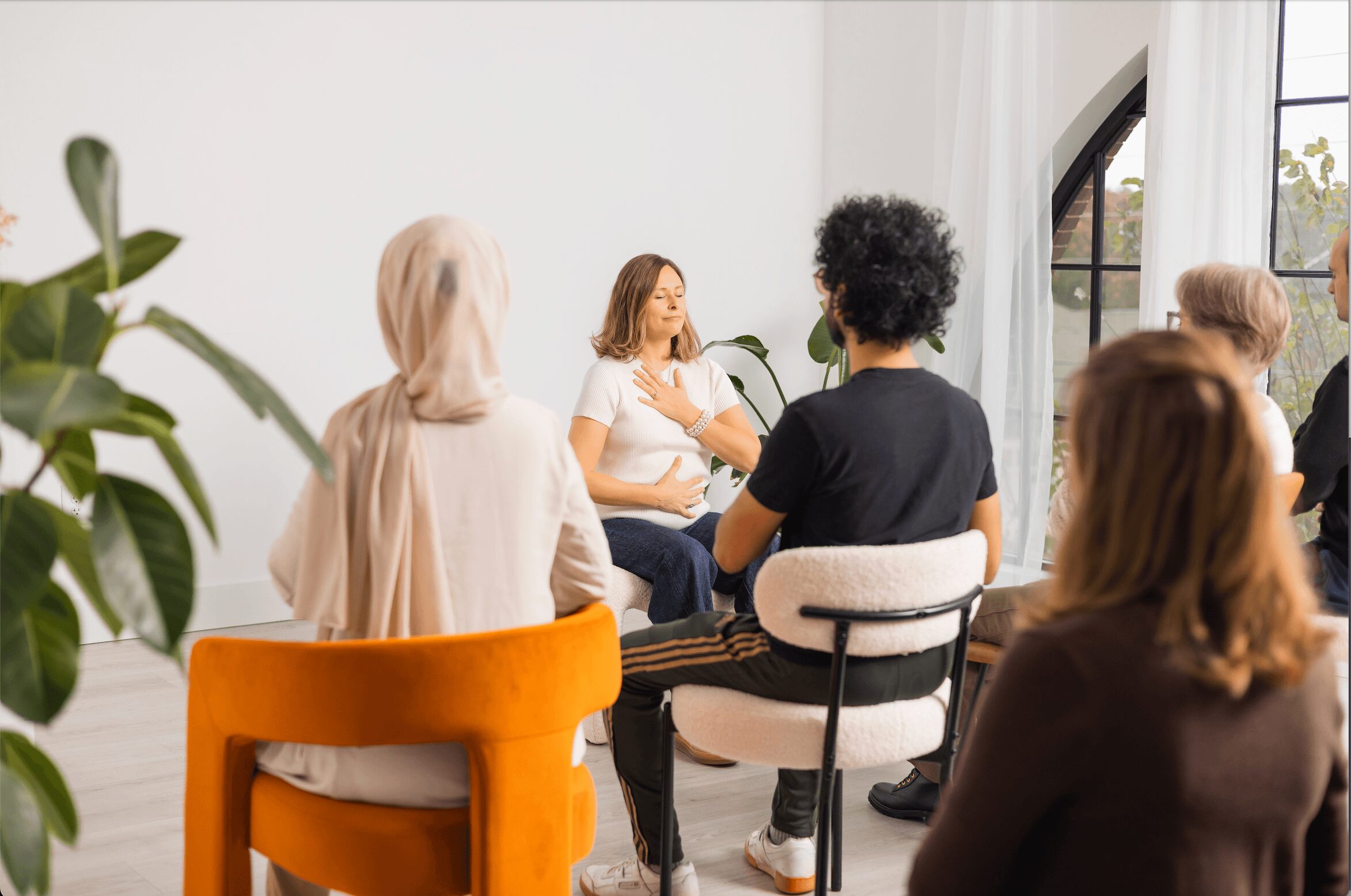 Group of people sitting in a circle during a discussion in a bright room with large window and plants.