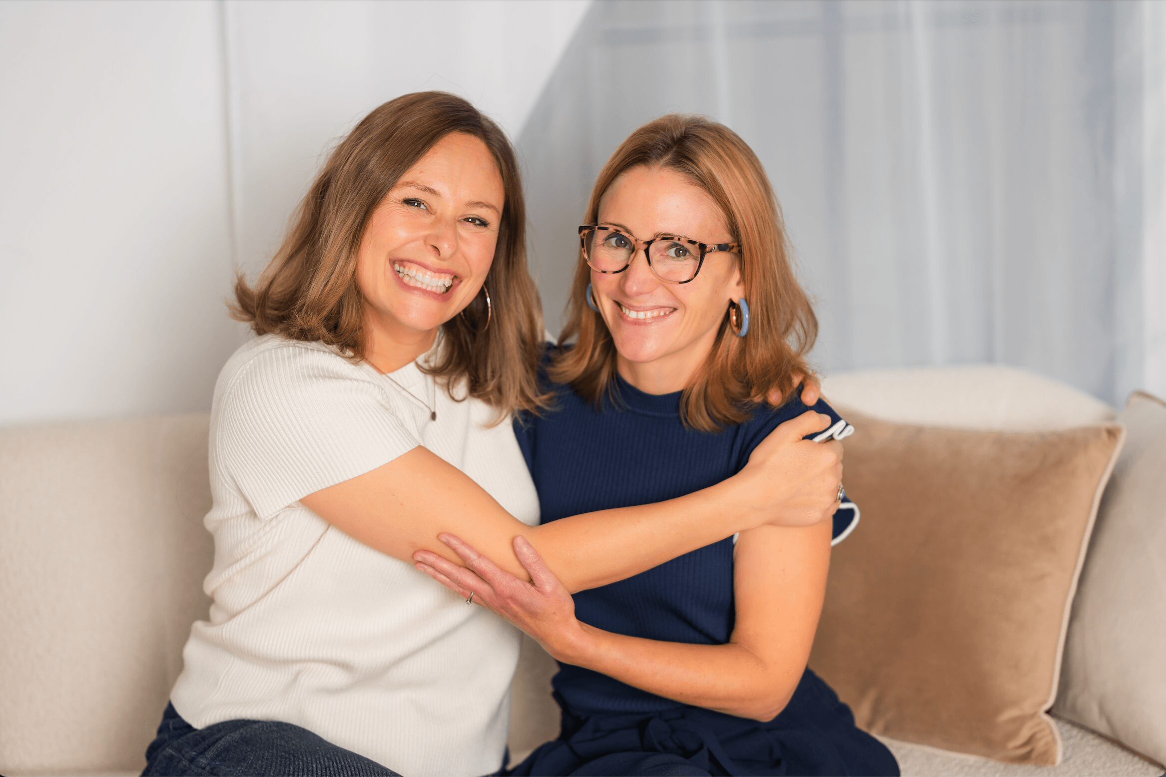 Two women sitting on a sofa, smiling and hugging each other, in a bright room with curtains.