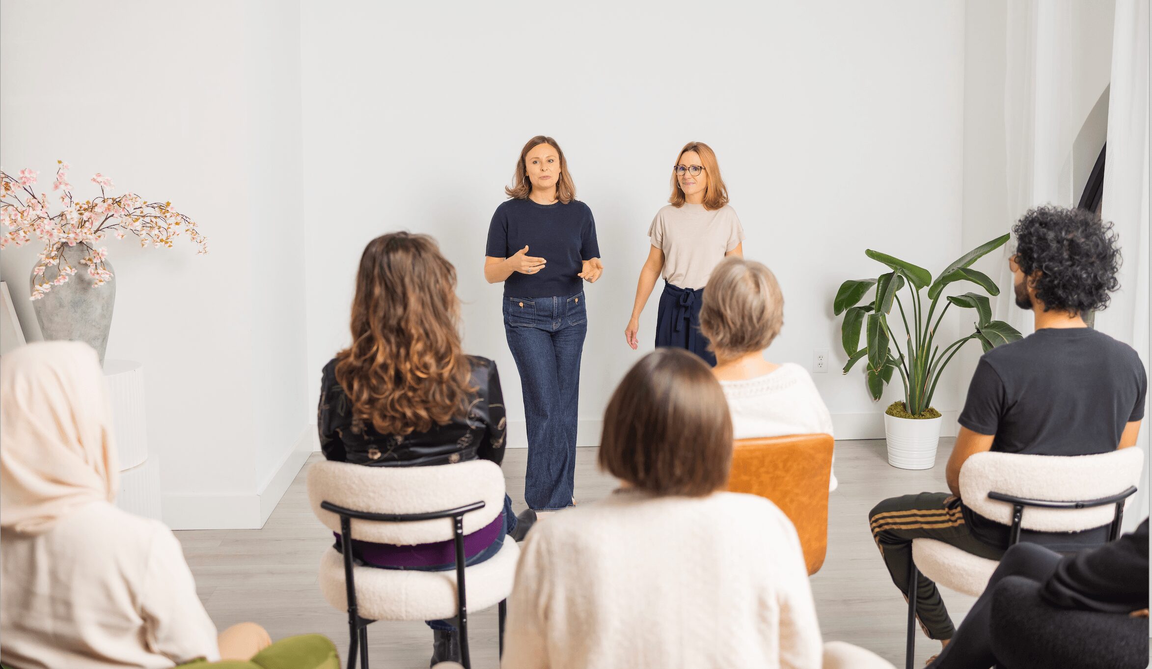 Two women stand in front of a seated audience in a bright room with white walls and large plants.