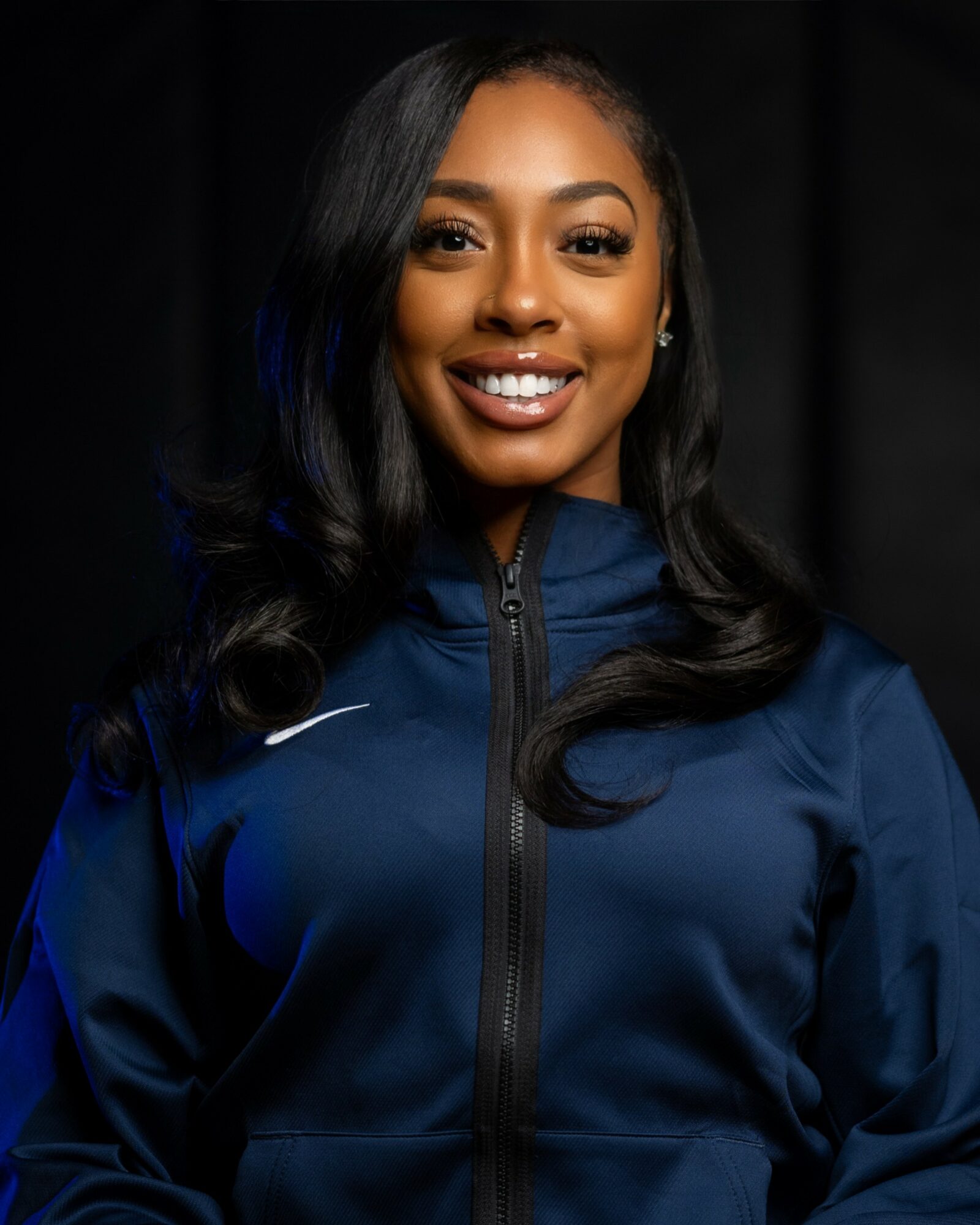 Smiling woman with long black hair wearing a blue sports jacket, dark background.