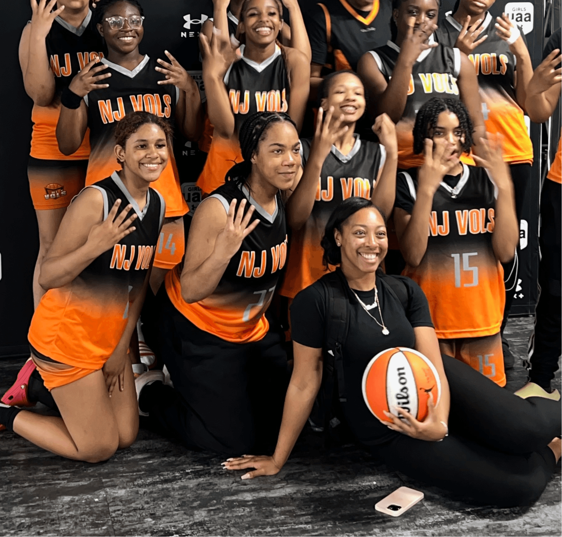 Group of female volleyball players in black and orange uniforms, some kneeling and some standing, holding a volleyball.