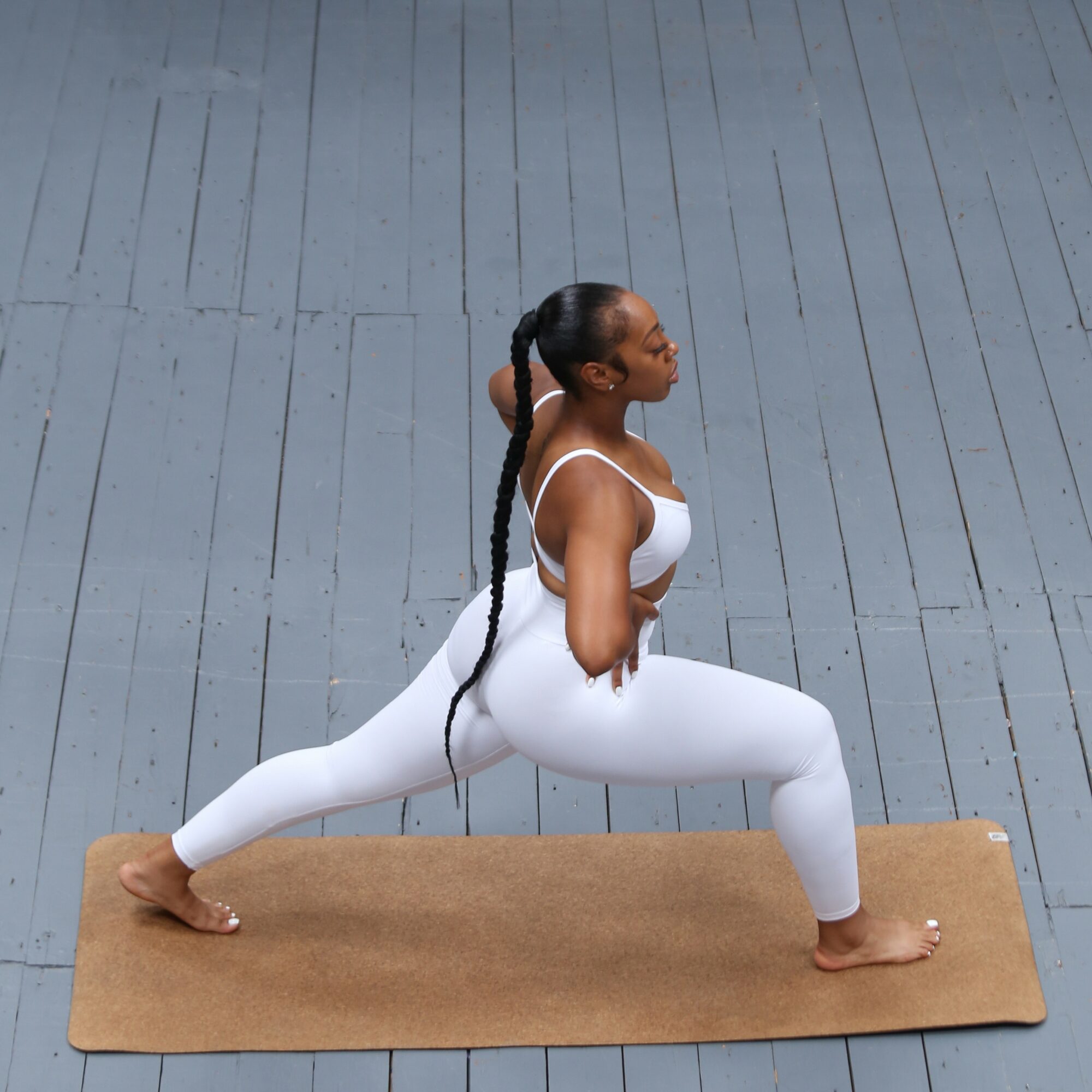 Woman practicing yoga in a lunge pose on a mat, wearing white workout clothes, on a wooden floor.