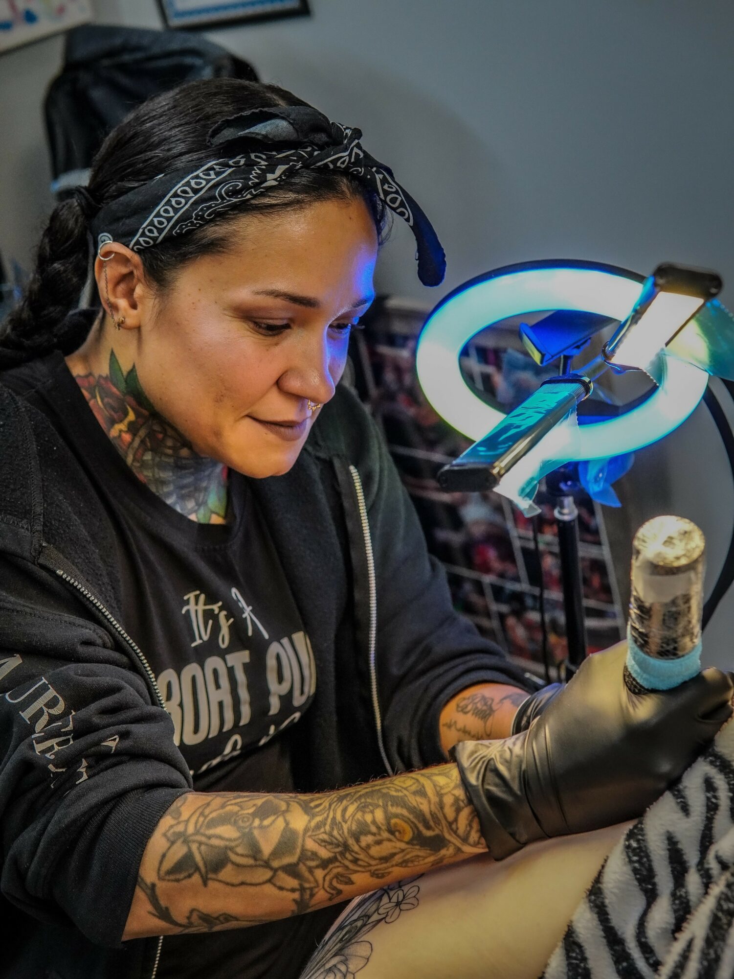 Tattooed woman with dark hair and bandana working on tattoo machine, focused, in tattoo studio.