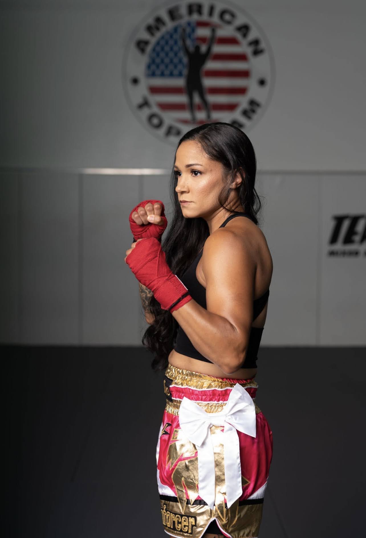 Female boxer in red gloves and shorts, standing in a gym with American flag logo in background.