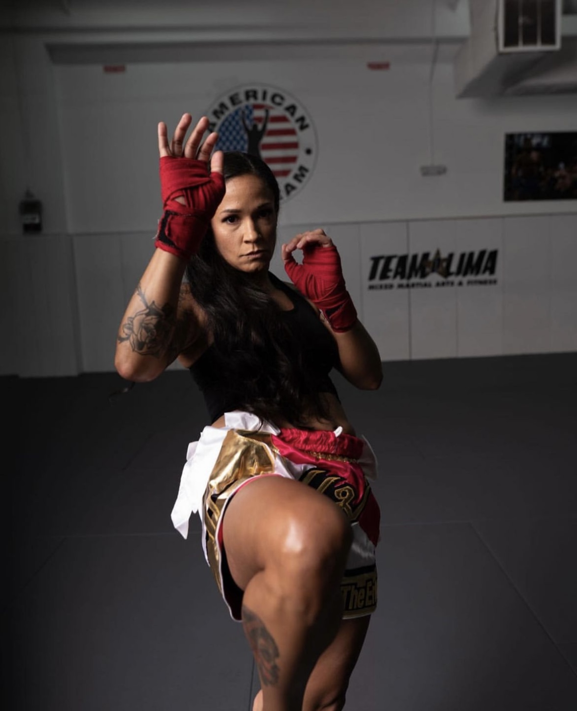 Female martial artist in fighting stance, wearing red gloves and shorts, in a gym with American flag and team logo.