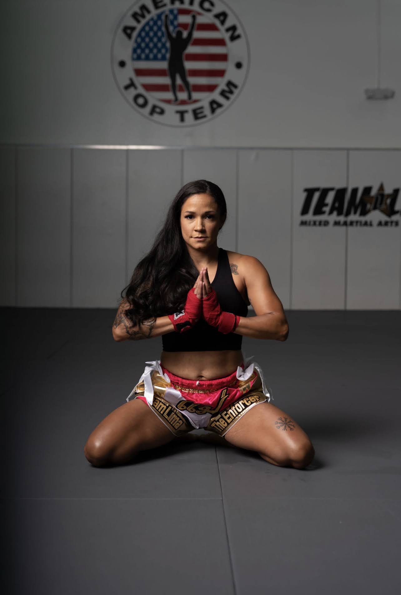 Woman in martial arts attire kneeling on mat with hands in prayer position, in training facility with American flag logo and banners.