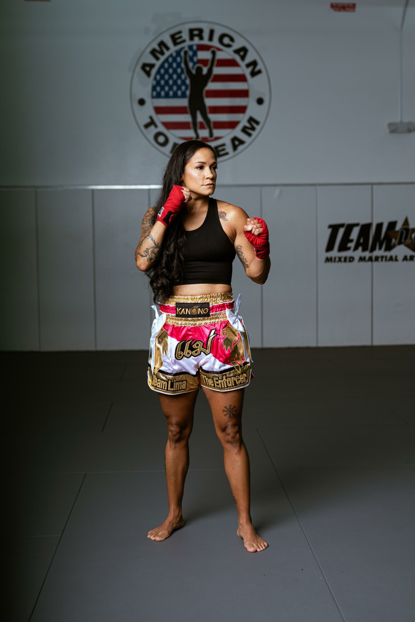 Female martial artist in boxing gloves and colorful shorts, standing in a gym with American Top Team logo in background.