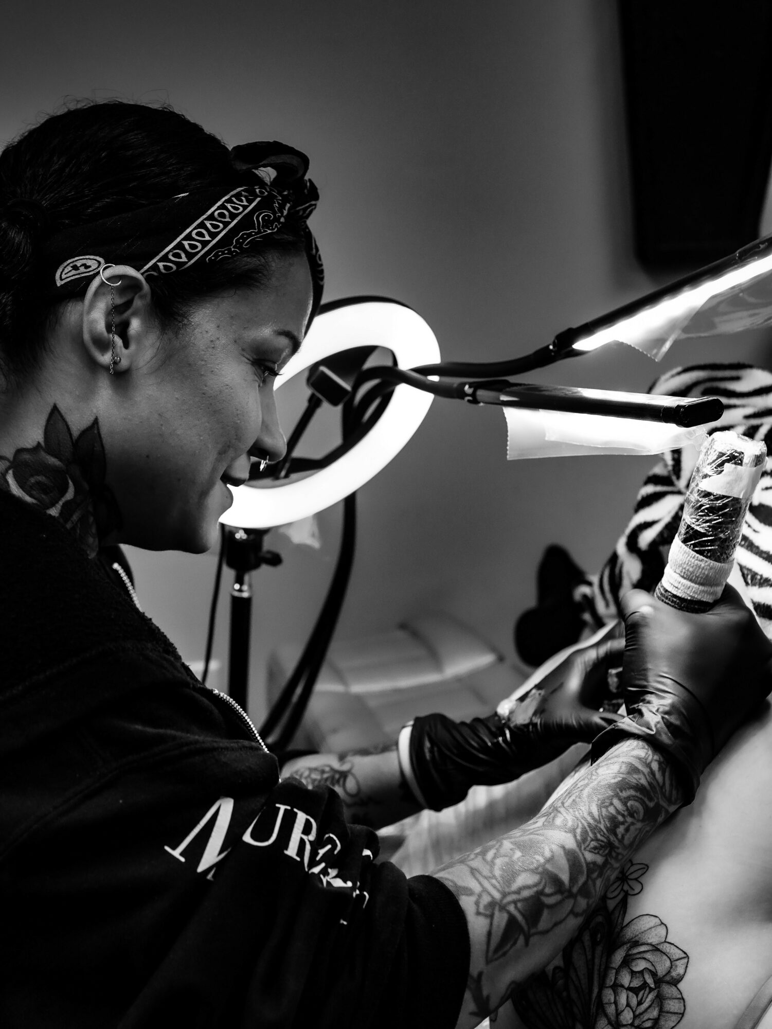 Tattoo artist working on a client's arm with tattoo equipment, in a studio setting, black and white image.