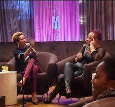 Two women sit on a sofa, engaged in conversation, with a microphone in one woman's hand, in a colorful room.