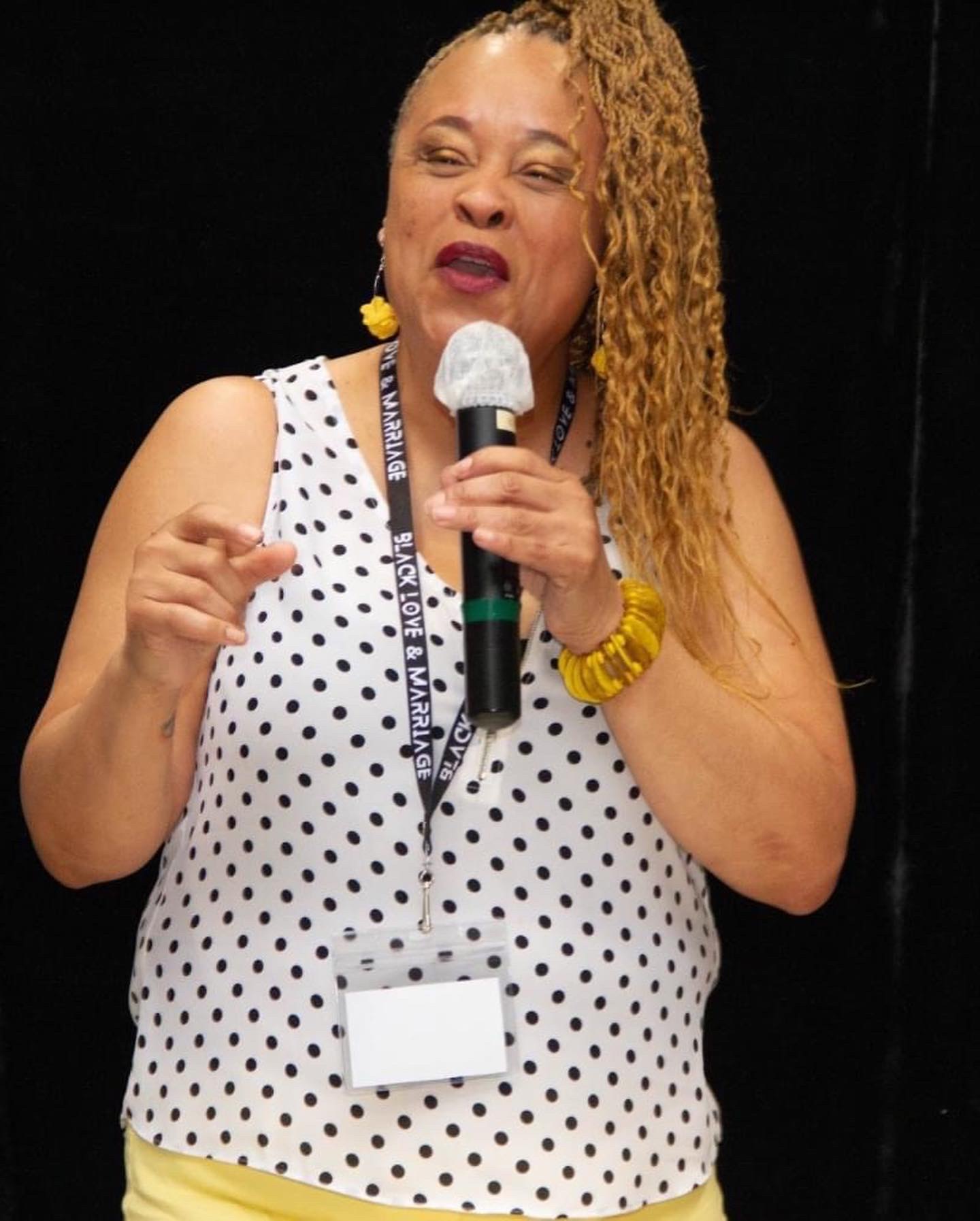 Woman speaking into a microphone, wearing a polka dot top, yellow accessories, and a lanyard, against a dark background.