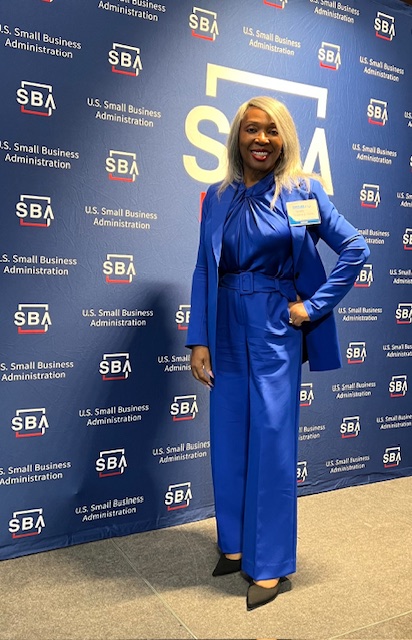 Woman in blue dress standing in front of SBA backdrop, smiling, with hand on hip.