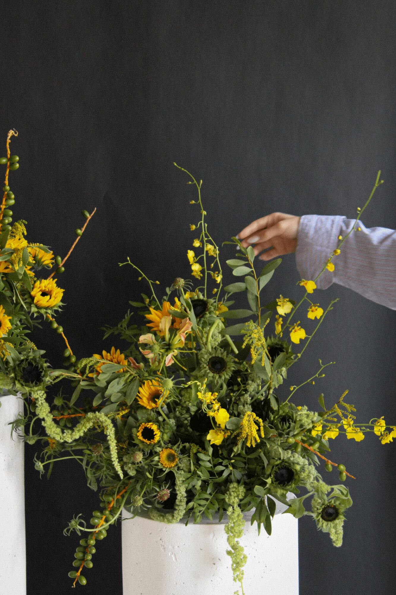 Person's hand reaching into a large flower arrangement with sunflowers and yellow flowers in a white container, dark background.