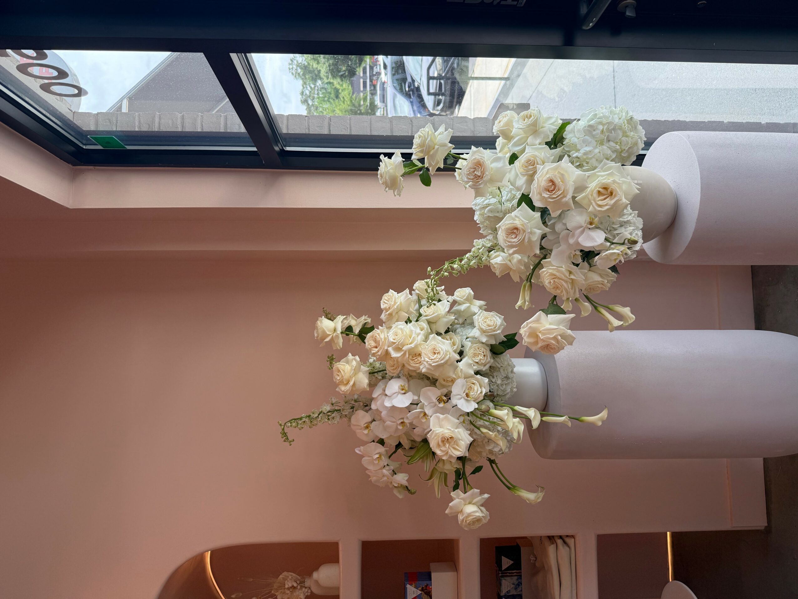 Two white vases with white and pale pink flowers on a windowsill, with a window showing outdoor scenery.