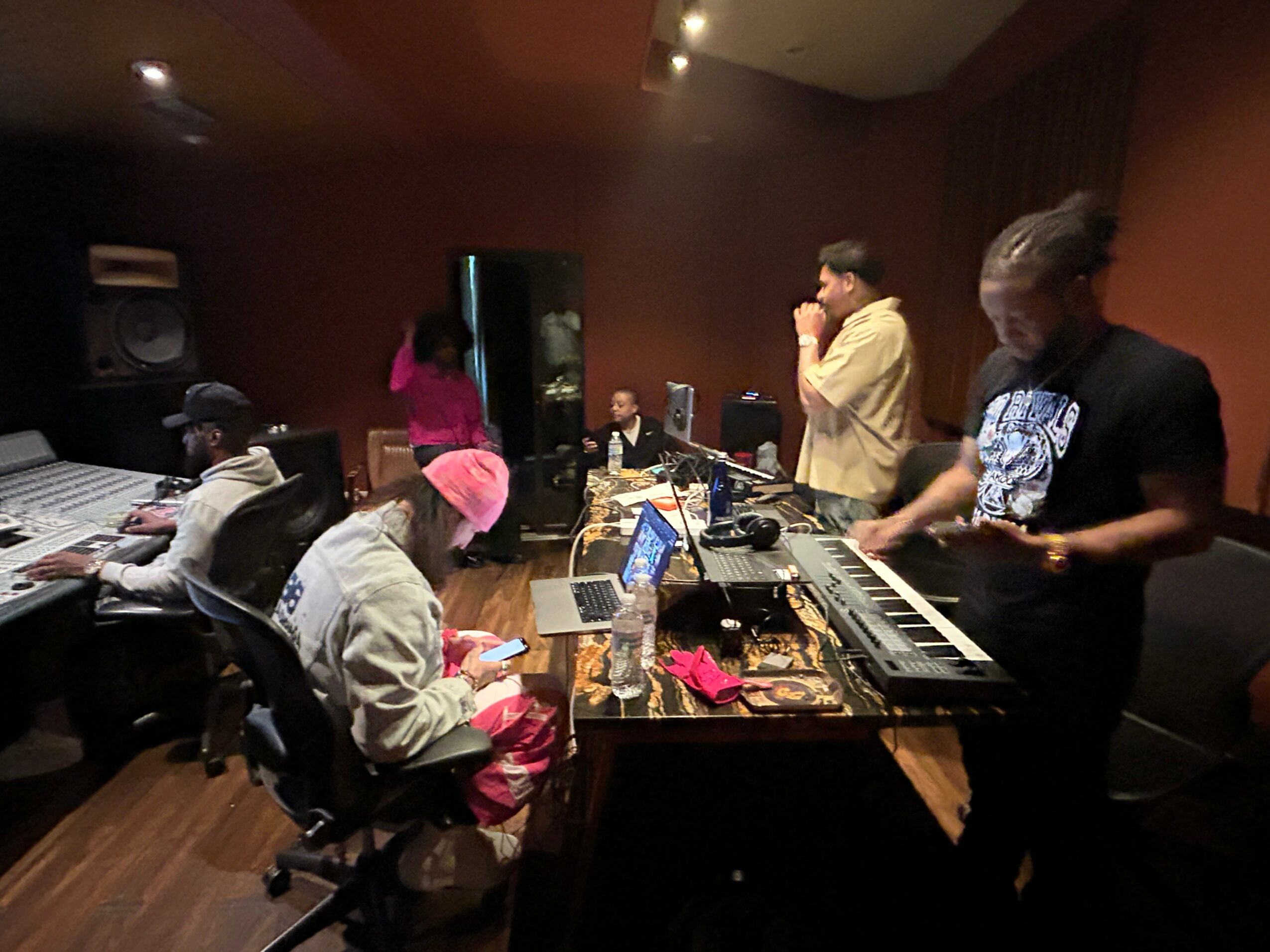 People in a music studio working with equipment and instruments, some sitting and some standing, dimly lit room.